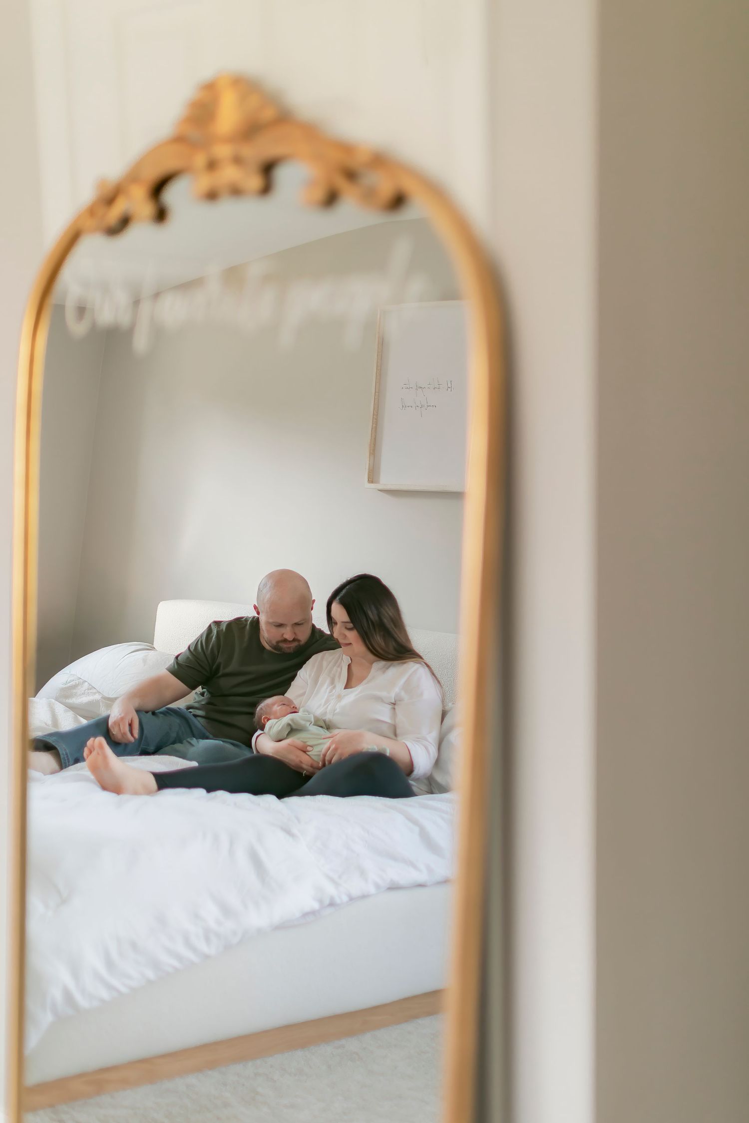 A couple sits on a white bed holding their newborn baby, reflected in an ornate gold-framed mirror.
