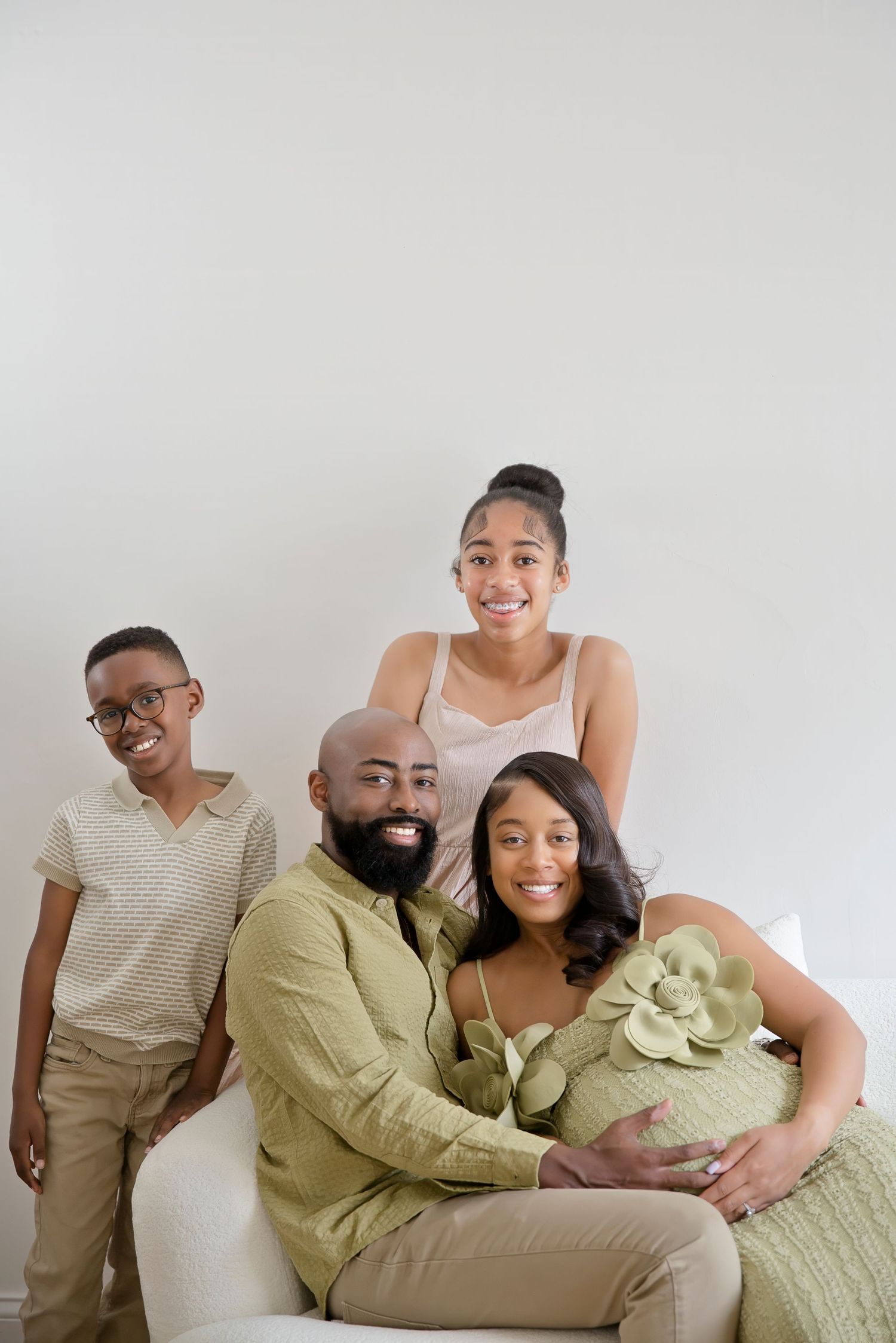 A joyful Black family poses together, with a pregnant woman in a green floral dress seated, surrounded by her husband and two children.