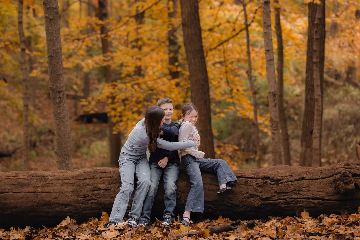 Family of three sitting on a fallen log in an autumn forest with golden foliage.