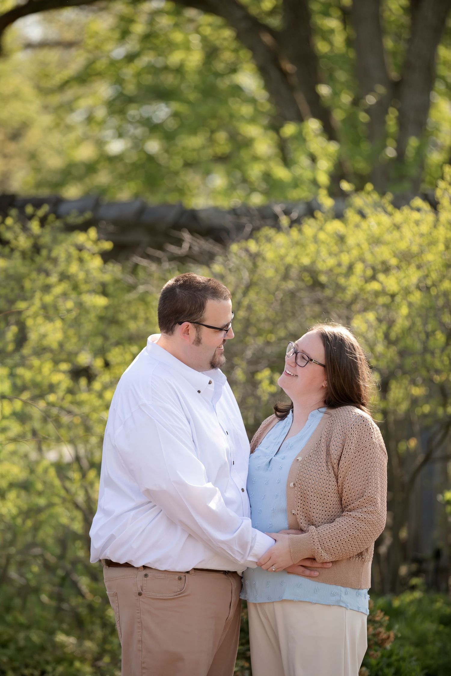 Couple holding hands and gazing at each other outdoors among spring greenery, wearing casual attire for an engagement photo.