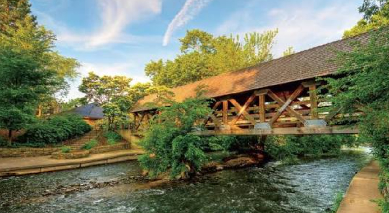 A scenic covered wooden bridge spans a gentle stream surrounded by lush green trees in a tranquil park setting.