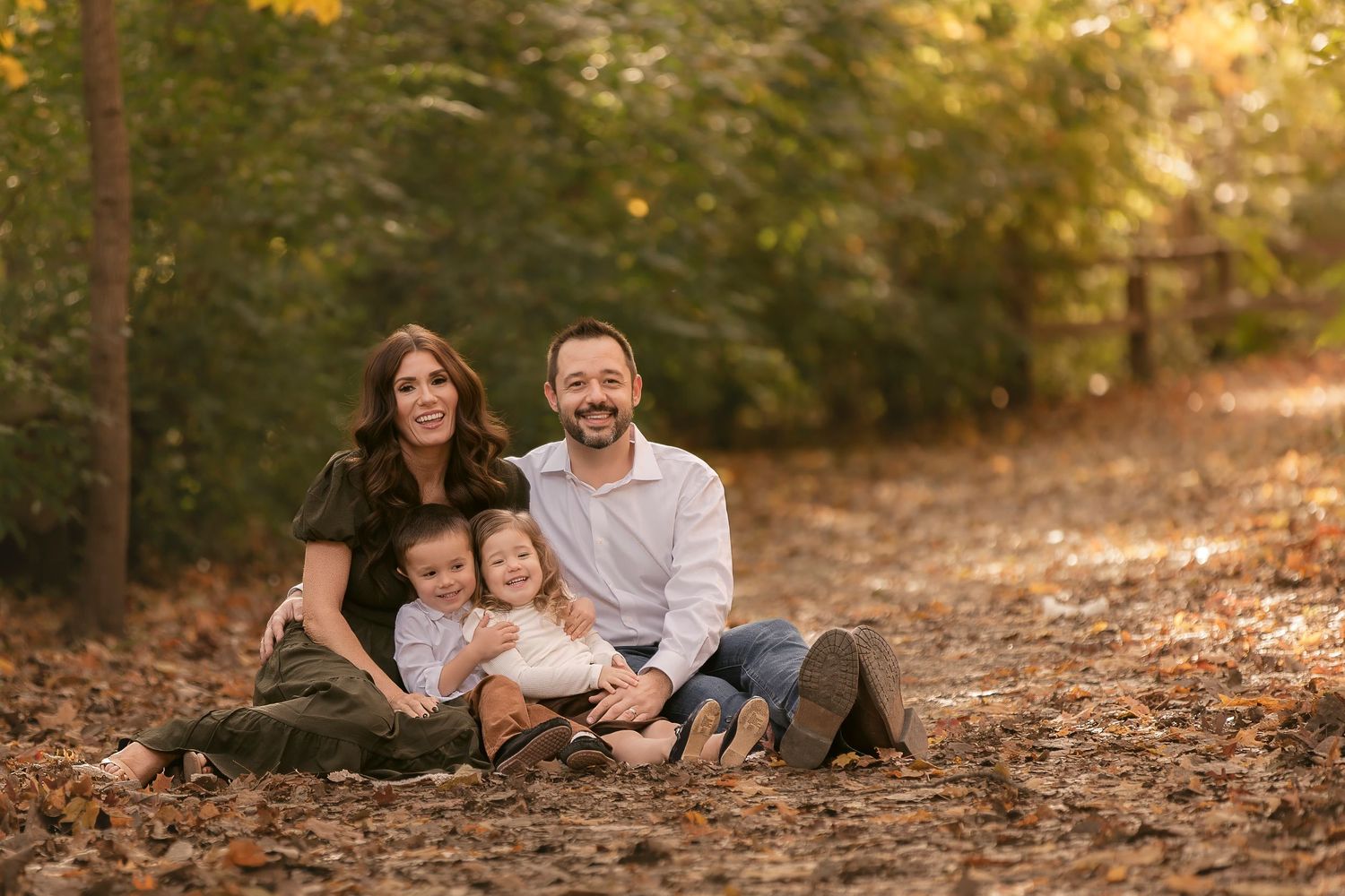 Family of four smiling together on a leaf-covered path surrounded by autumn trees in a warm outdoor portrait.