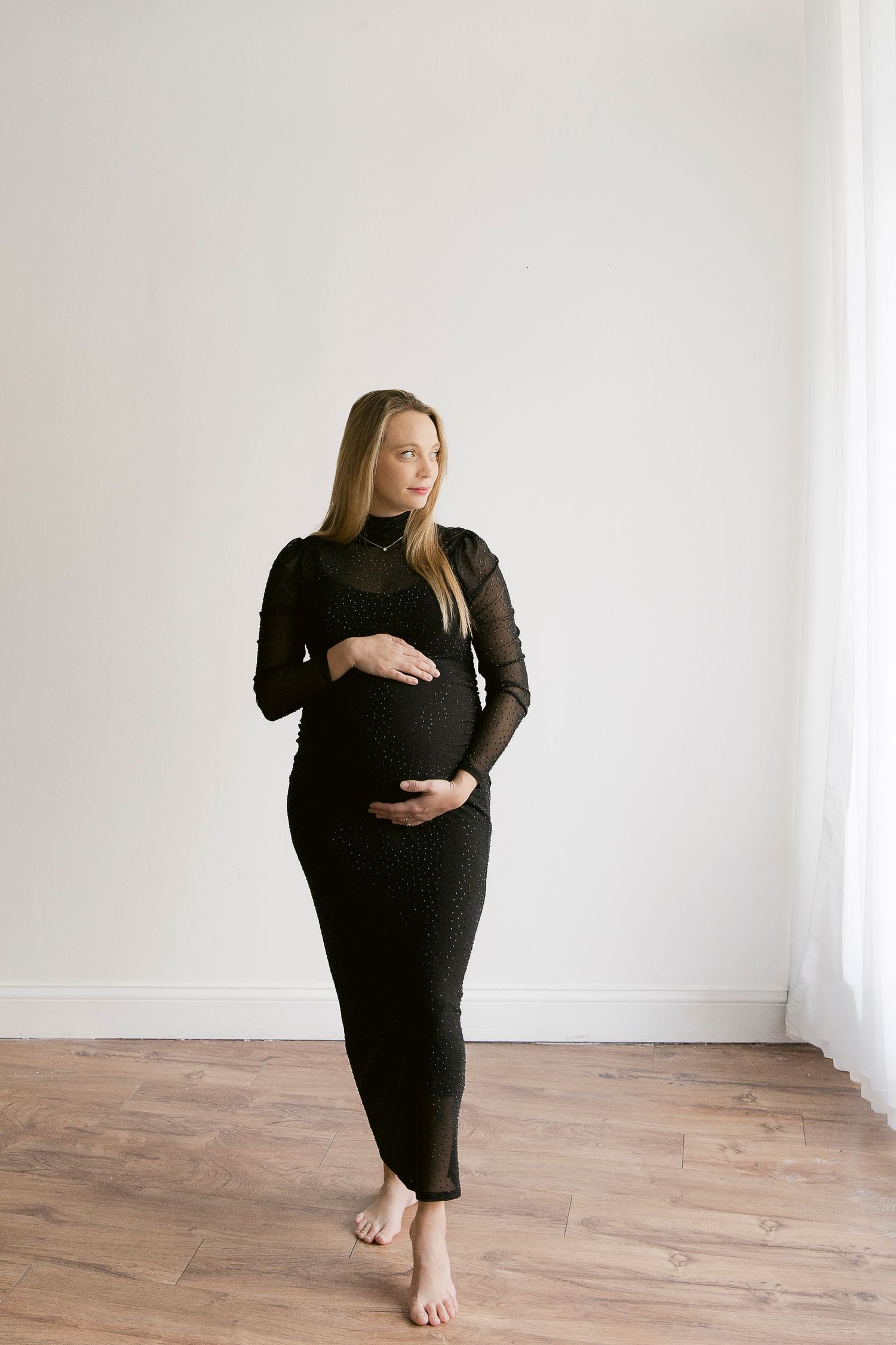 Pregnant woman in black dress poses gracefully against white wall during maternity photography session.