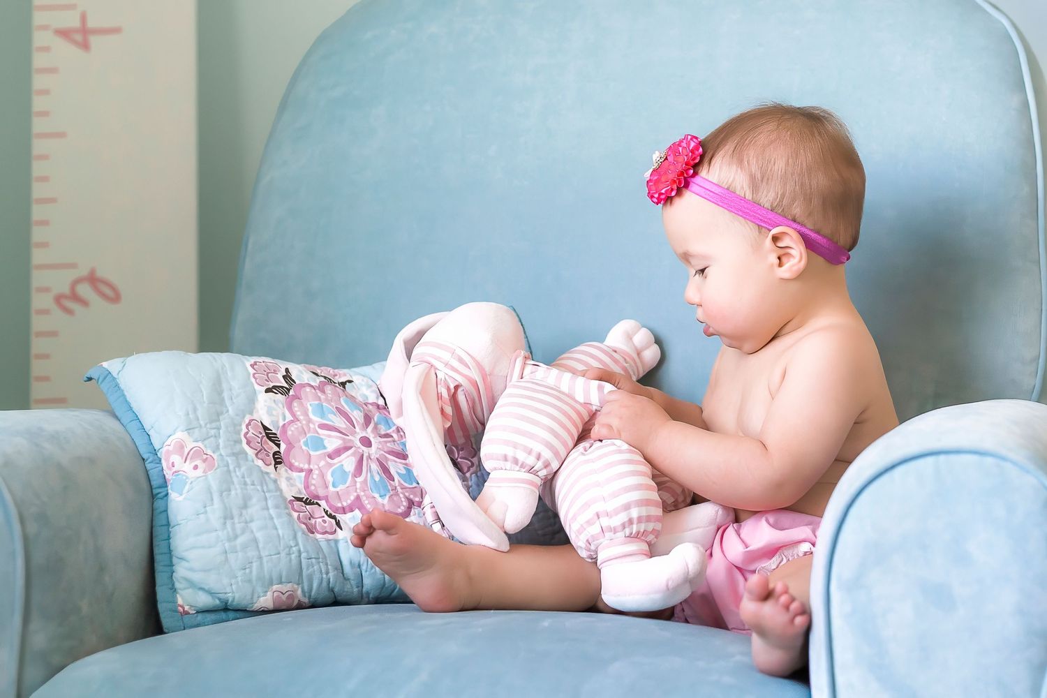 Small baby wearing pink headband sits on blue chair while putting on striped leg warmers.