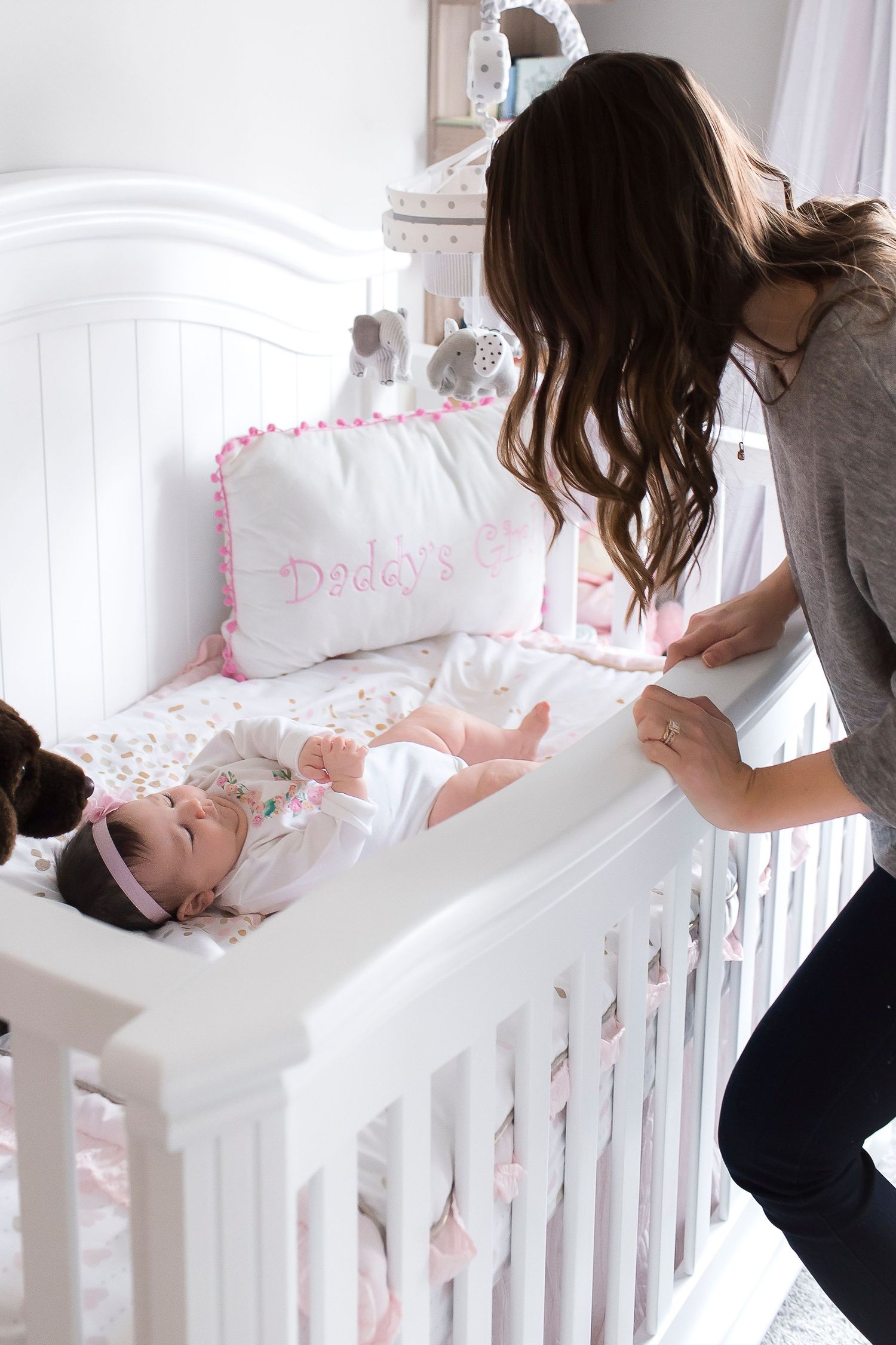 Baby lying in white wooden crib with pink bedding while being tended to in a bright nursery room.