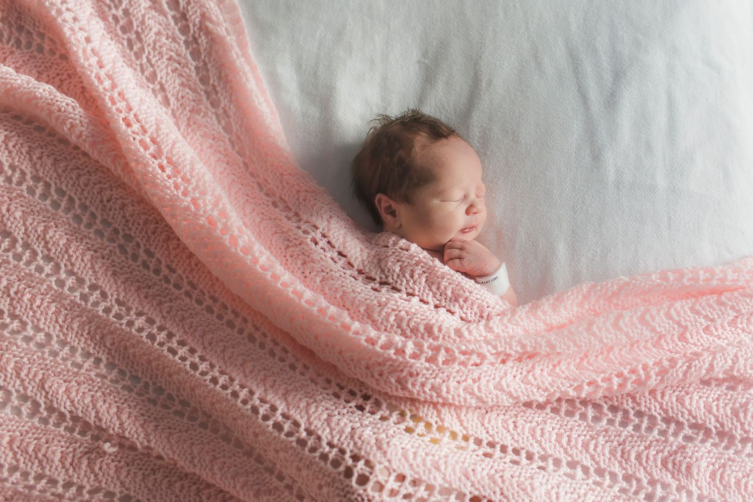 Newborn wrapped in soft pink knitted blanket resting on white bedding.
