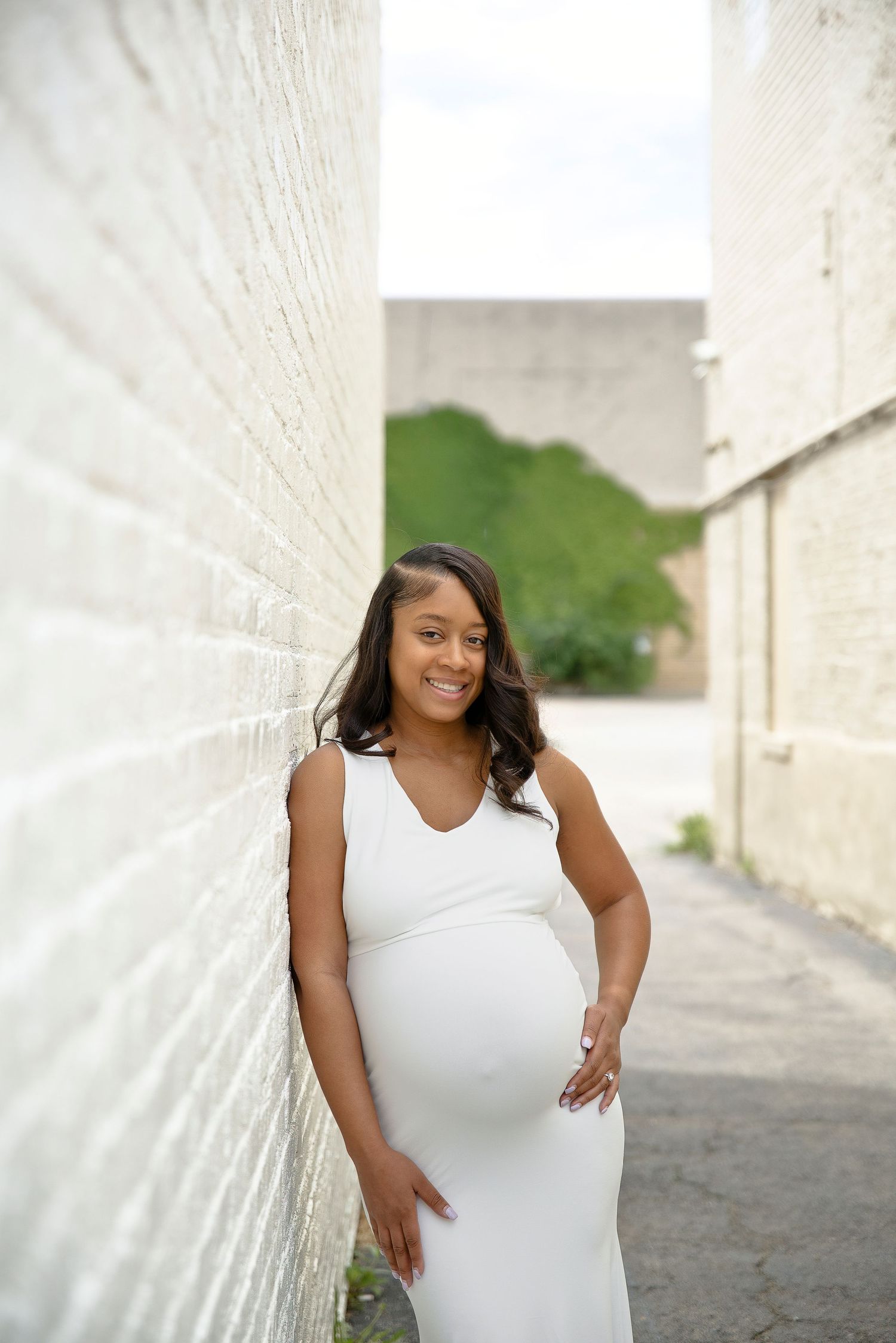 Smiling pregnant woman in white dress leaning against white brick wall in outdoor alley.