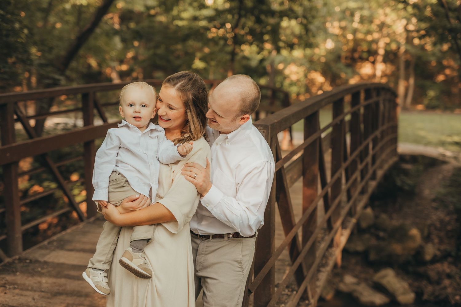 A happy family of three poses on a wooden bridge surrounded by lush green trees in a warm outdoor setting.