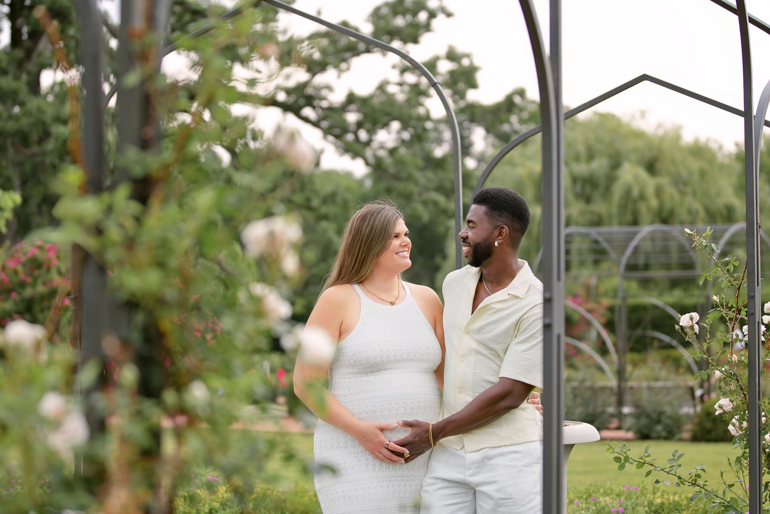 Interracial couple expecting a baby poses in a lush garden under a metal arch surrounded by roses.