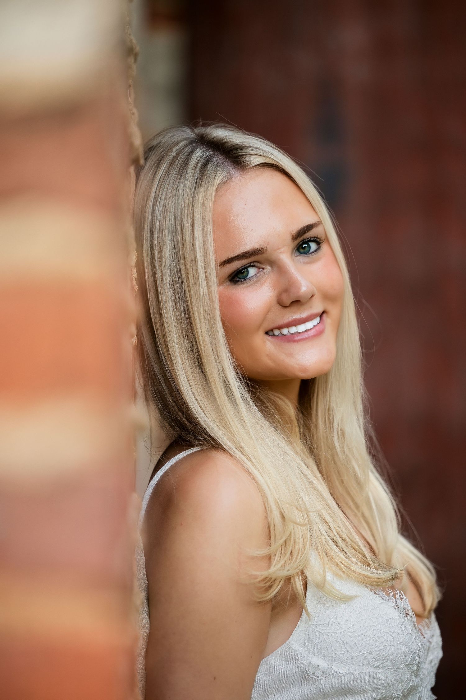 Young blonde woman with blue eyes smiling warmly, leaning against a red brick wall outdoors.