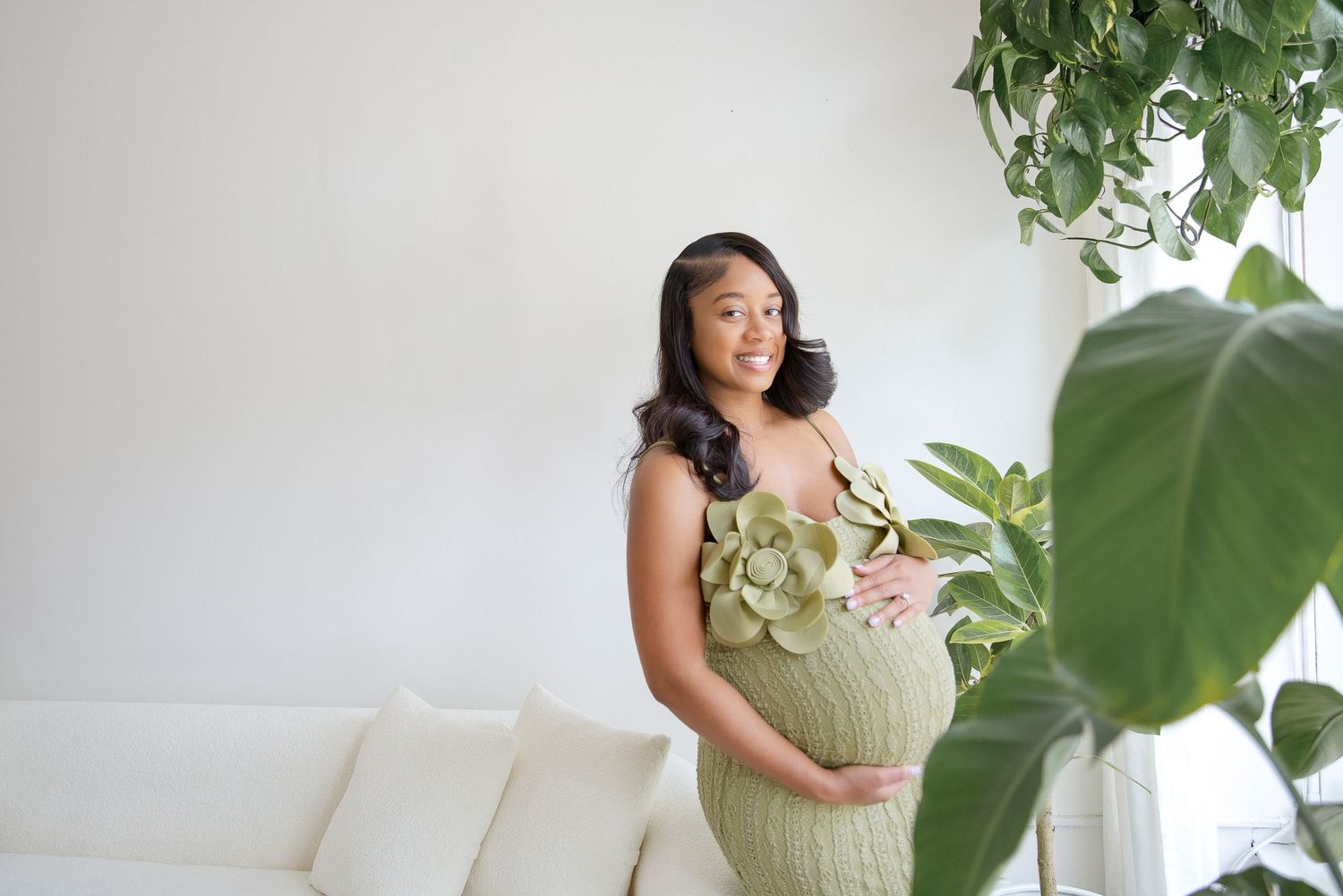 Pregnant woman in green floral dress holding baby bump indoors surrounded by lush tropical plants.