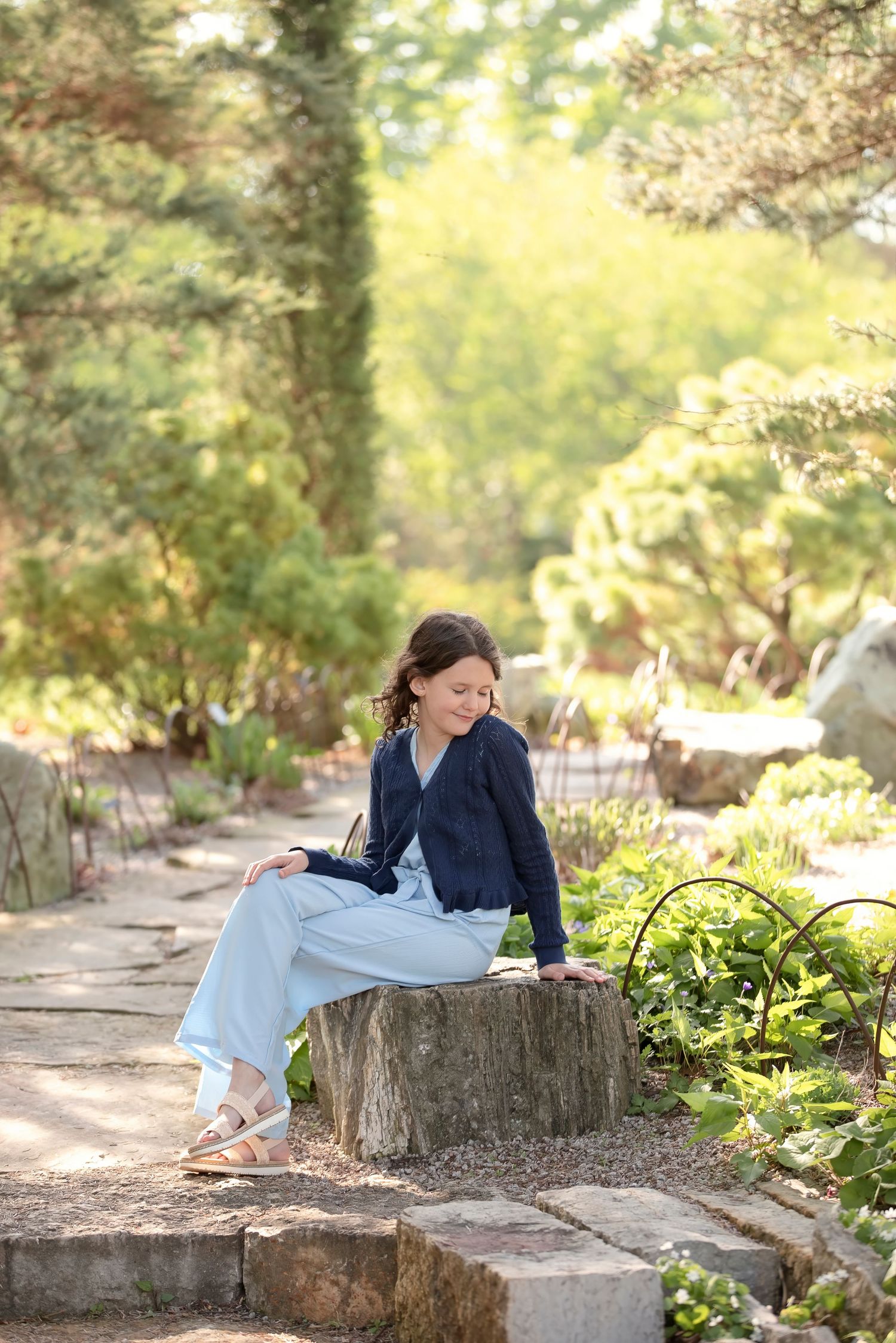 A smiling woman in navy cardigan and light blue pants sits on a tree stump in a sunlit garden path.
