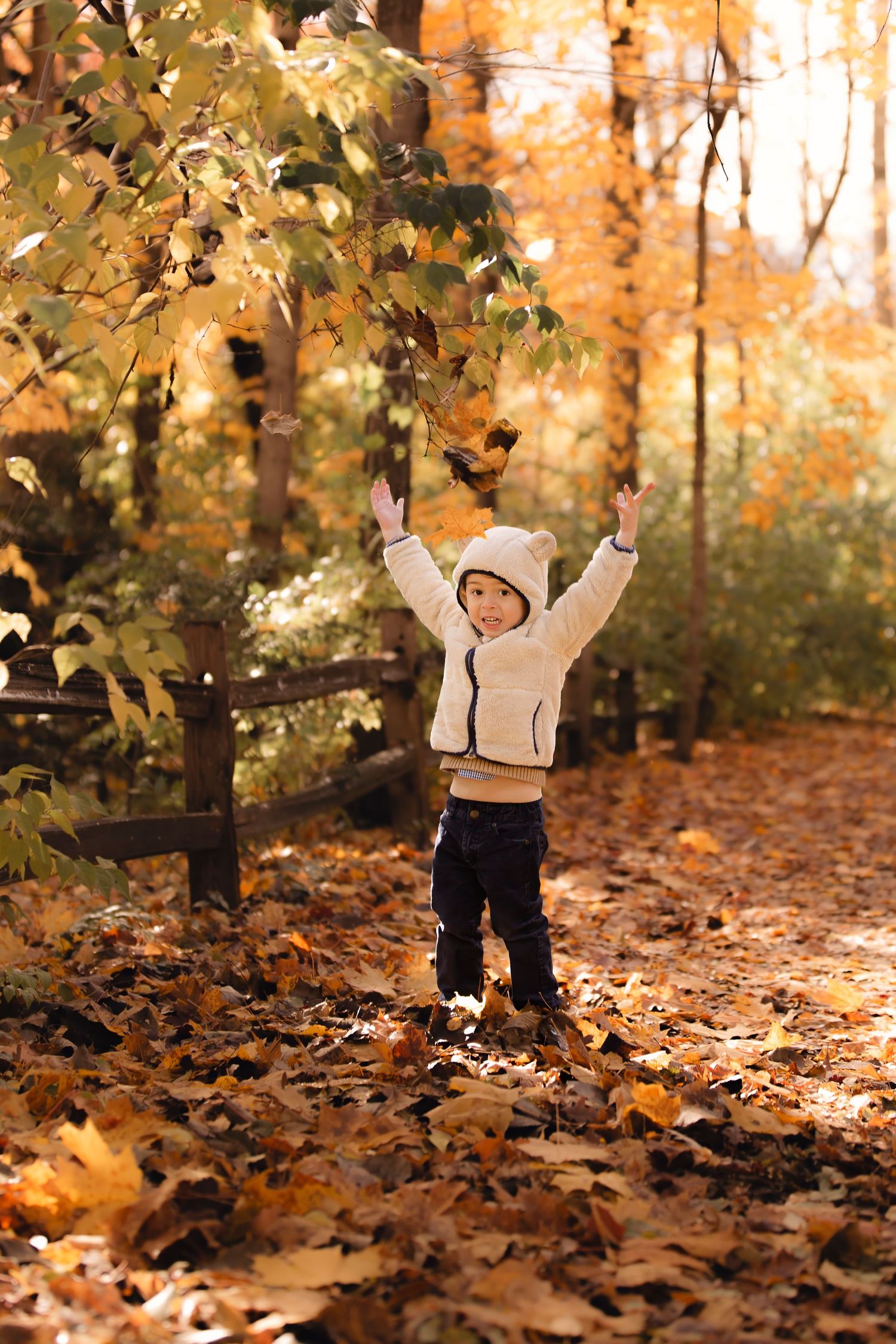 A child plays joyfully in fallen autumn leaves while wearing a cream sweater and dark pants in a golden forest setting.