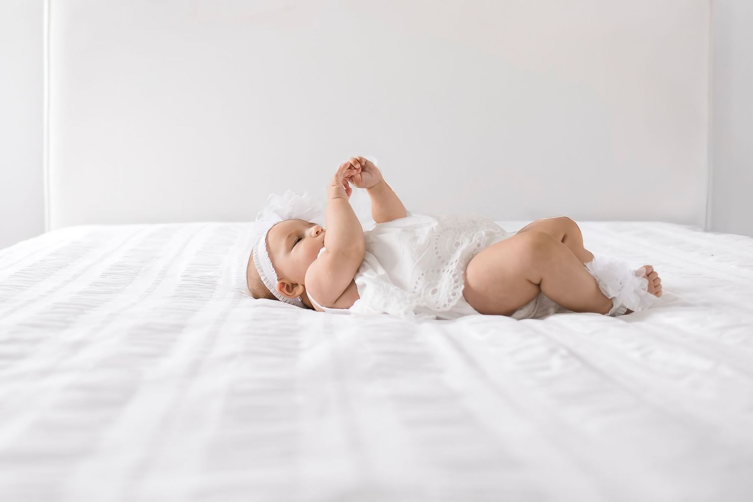 Baby wearing white onesie lies on white bedding in bright natural light.