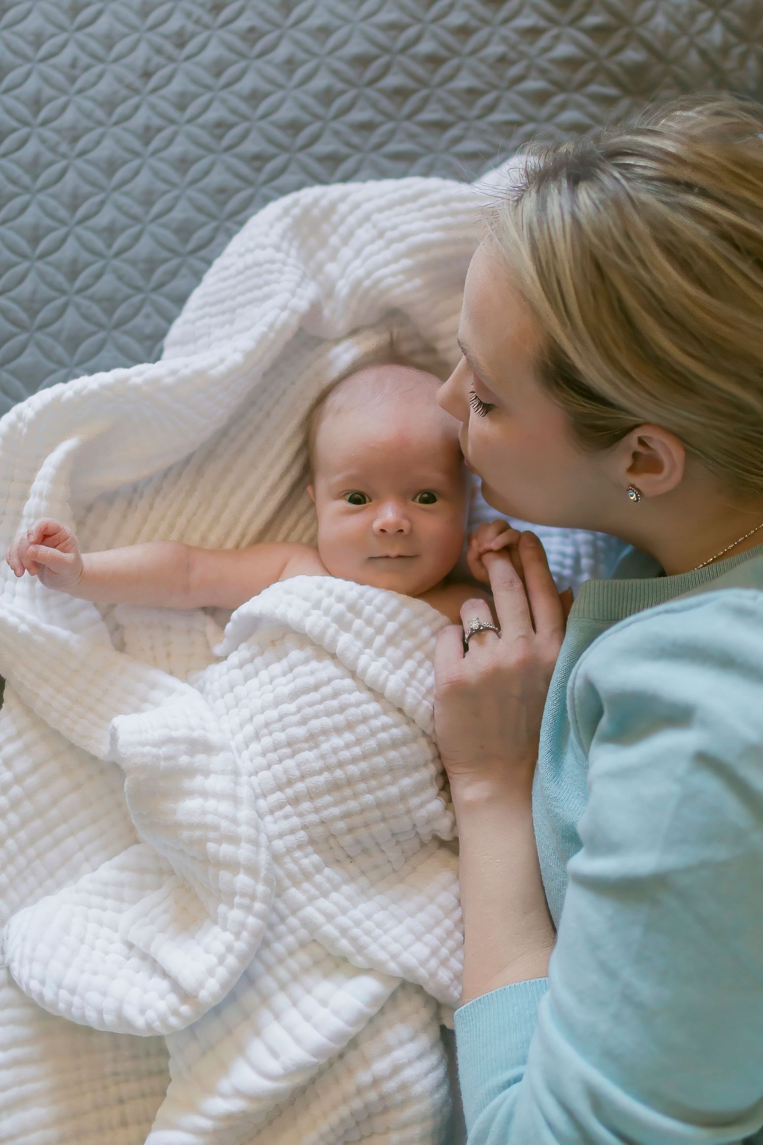 Mother in teal shirt kissing newborn baby wrapped in white knit blanket on gray bed.