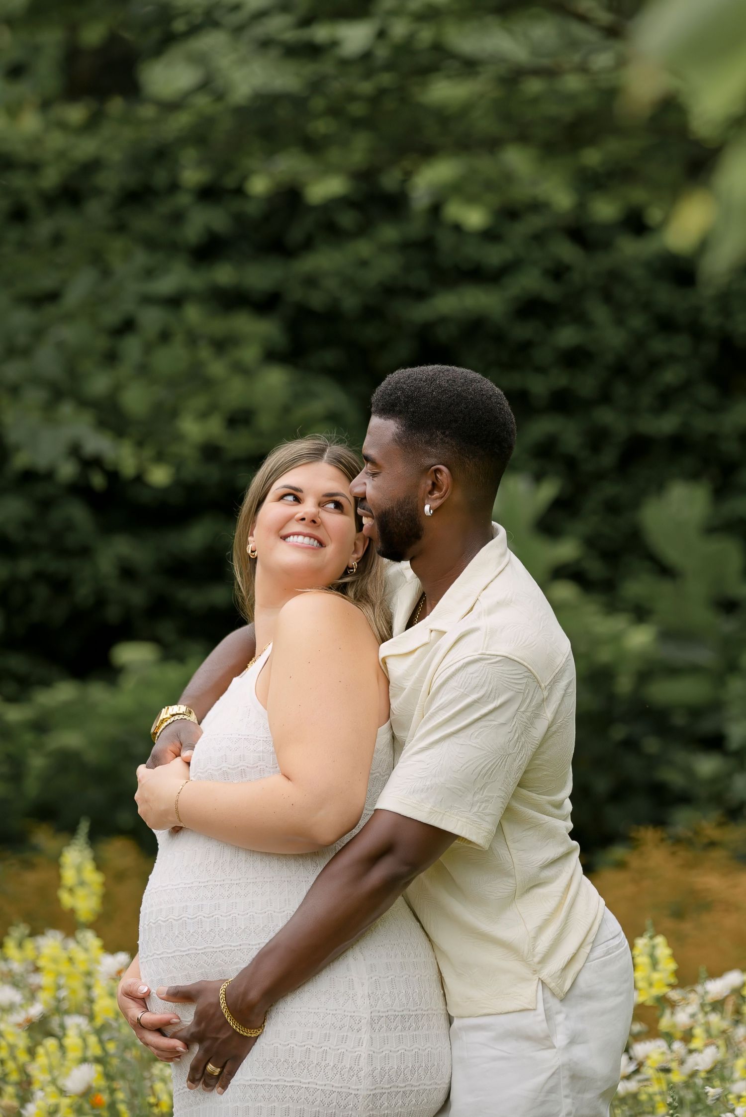 A couple embraces outdoors, the man kissing the pregnant woman's cheek amid lush green foliage.