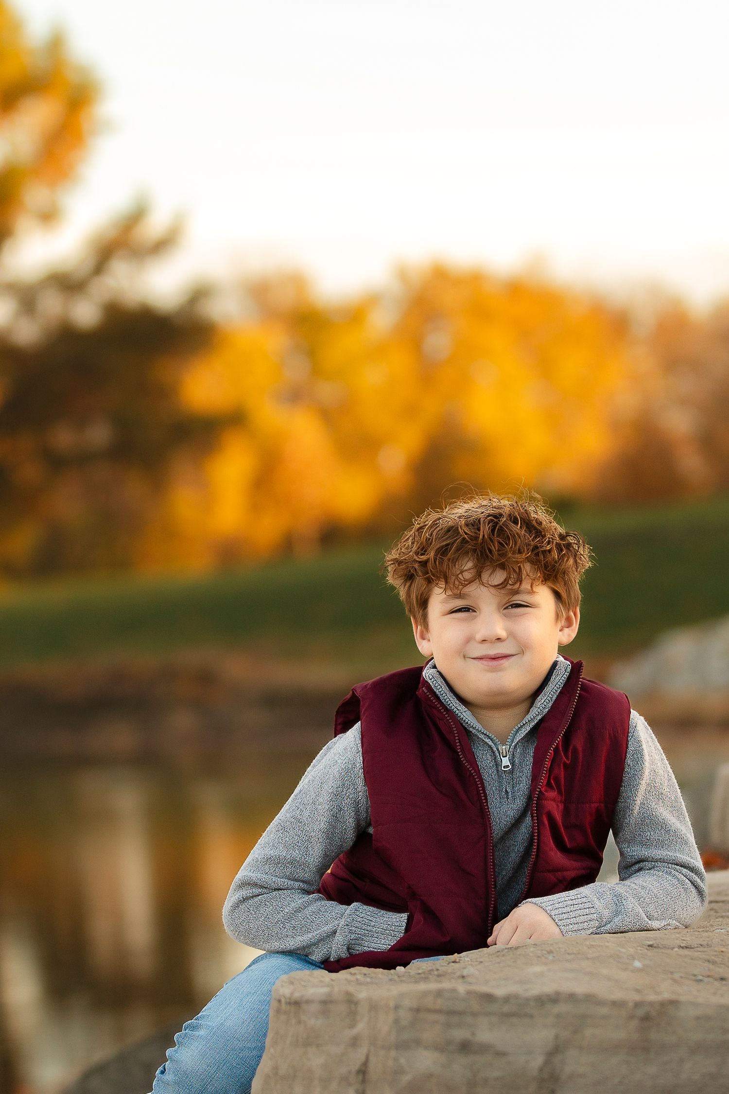 Child wearing burgundy vest and gray sweater sits by autumn pond with golden foliage in background.