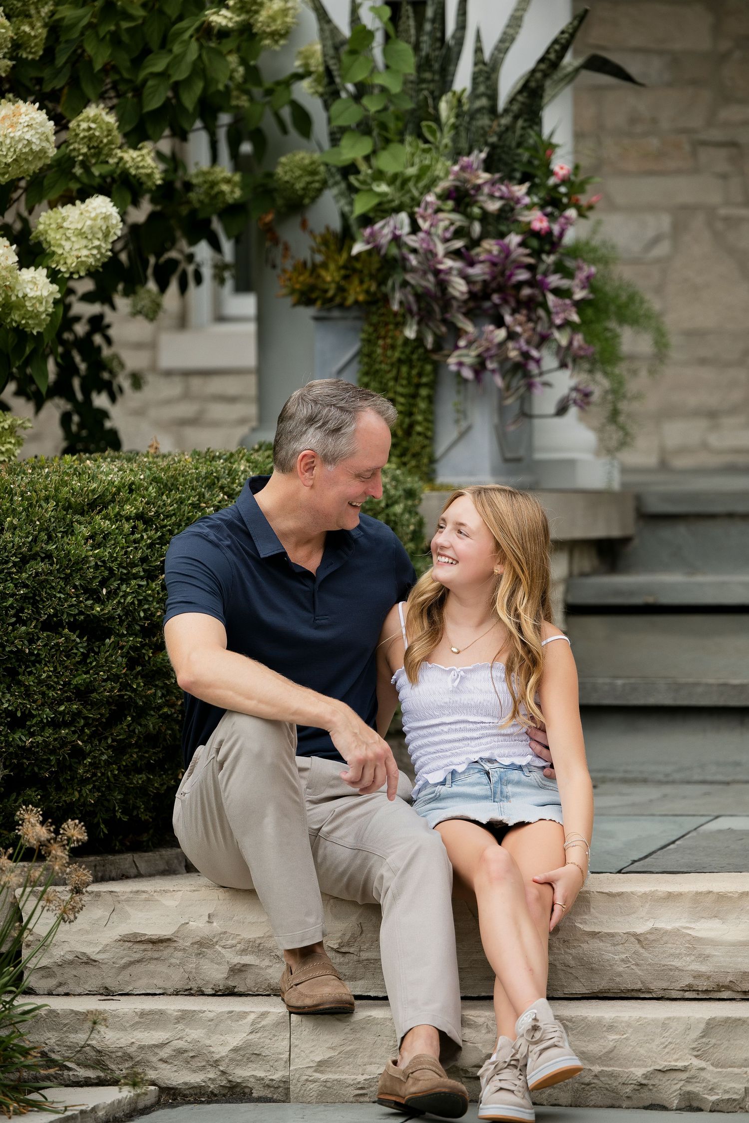 A man and teenage girl sit together on stone steps, smiling at each other, surrounded by lush garden flowers.
