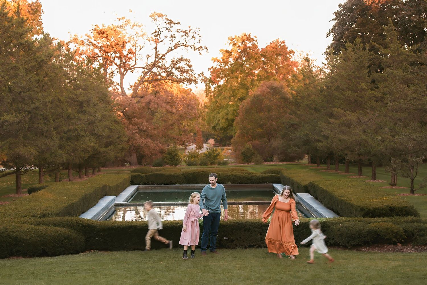 People in formal attire pose together in a scenic autumn park setting with golden foliage at sunset.