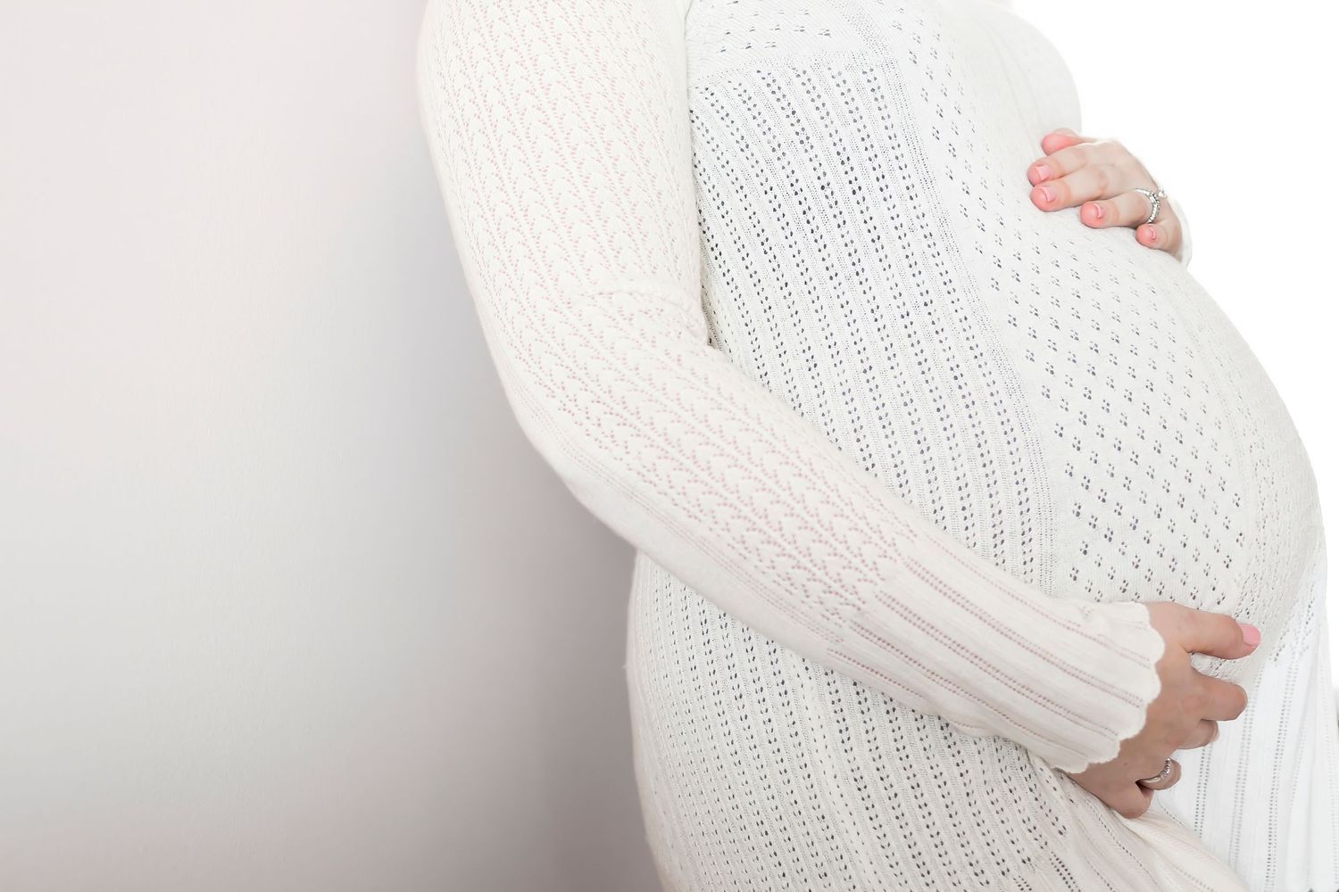 Pregnant woman in white knit sweater cradles her baby bump while standing against a light background.