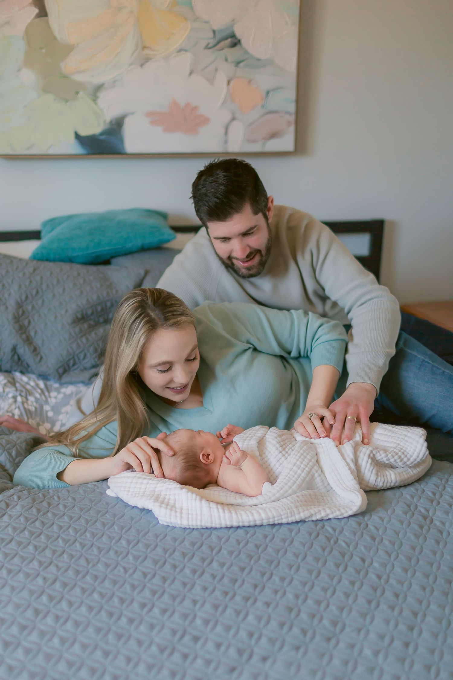 Happy parents smiling at their newborn baby lying on a bed with gray bedding in a cozy bedroom.