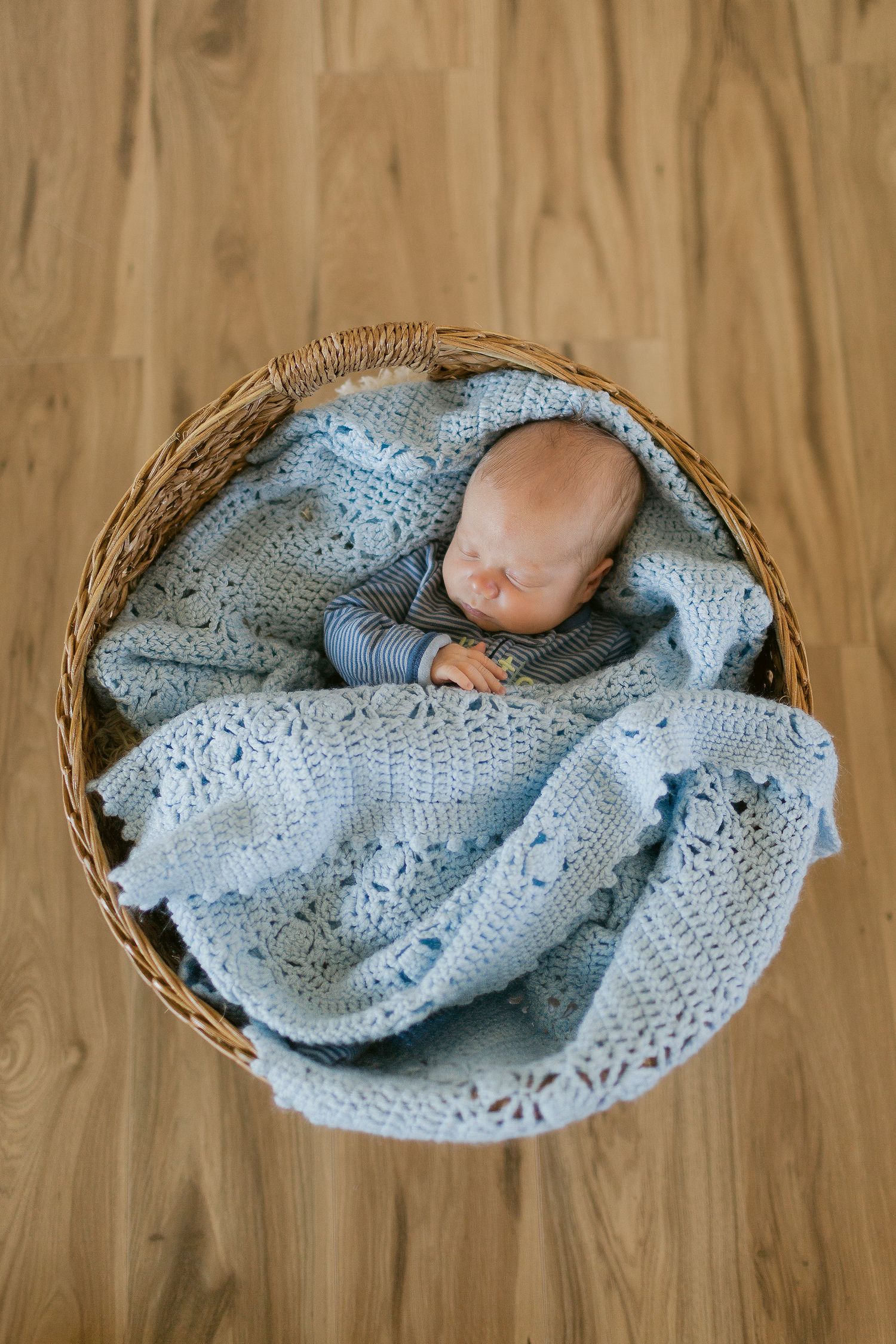 Newborn baby sleeping peacefully in a wicker basket wrapped in a soft blue crochet blanket on a wooden floor.