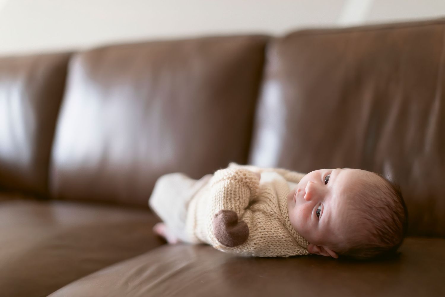 Newborn baby in knit outfit lying on brown leather sofa, looking up with wide eyes in cozy home setting.
