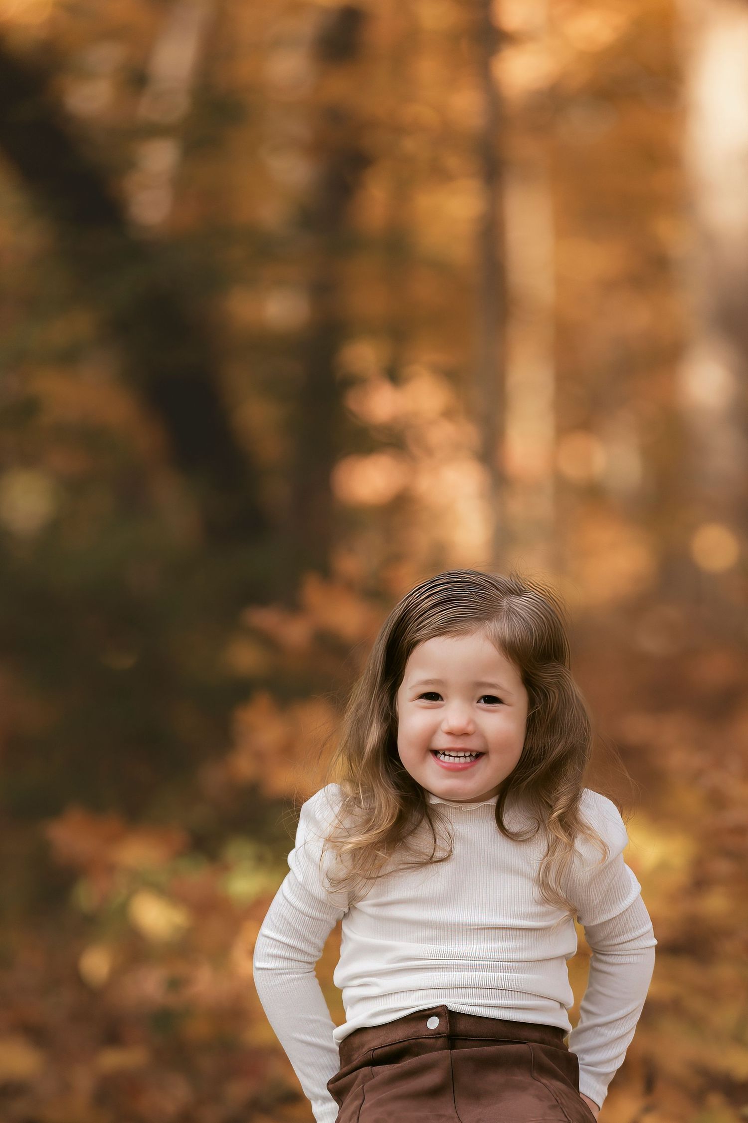 Smiling young girl in white turtleneck and brown skirt posing in autumn forest with golden fall foliage background.