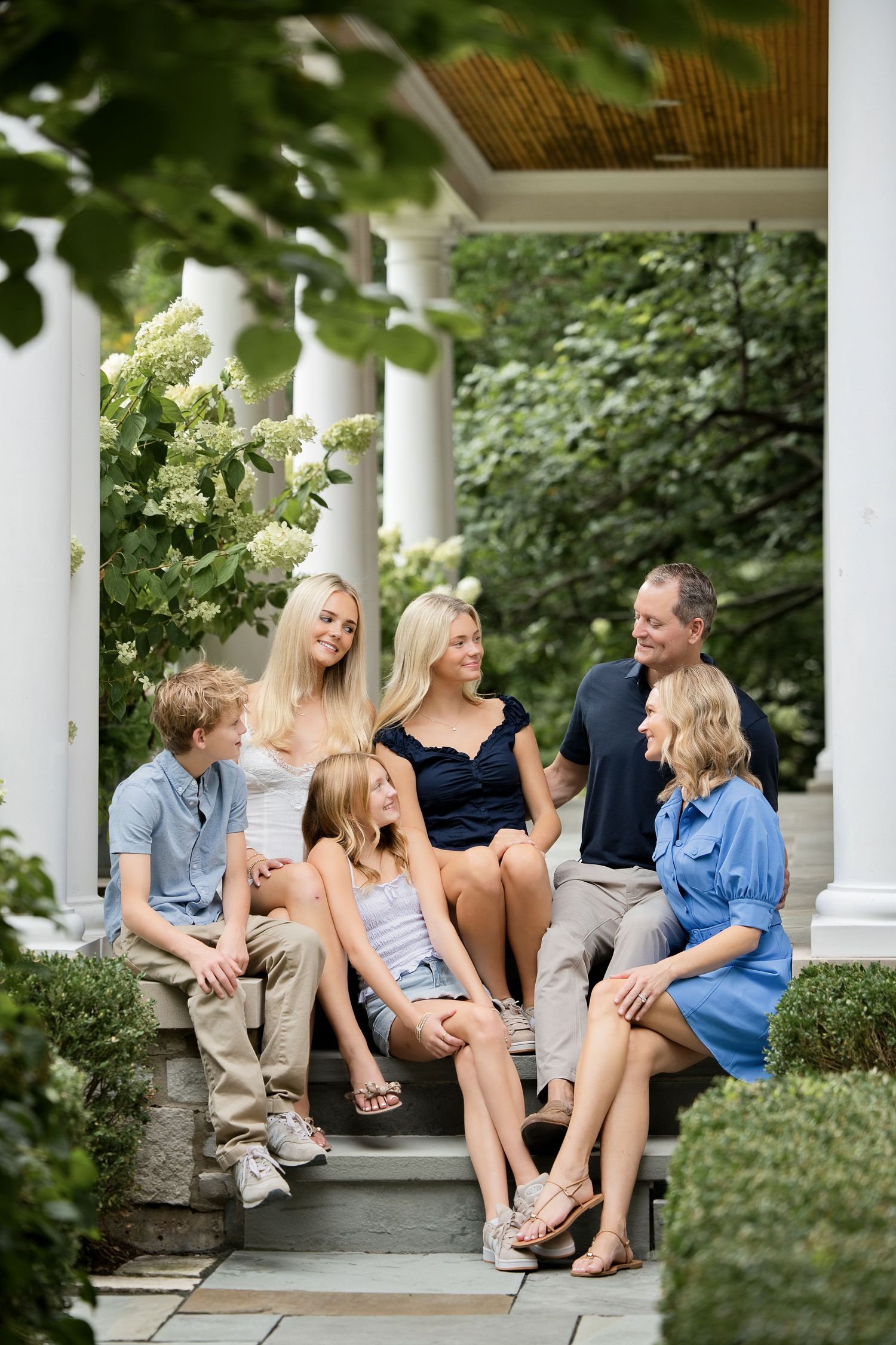 A happy family of six sits together on porch steps surrounded by white columns and blooming hydrangeas.