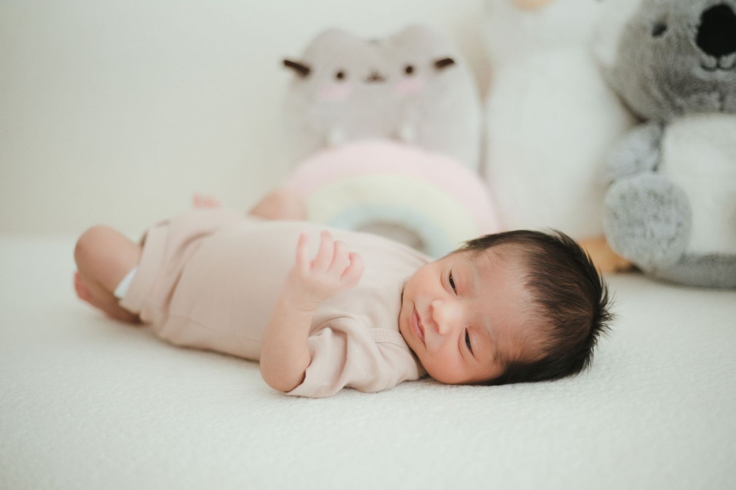 Newborn baby sleeping peacefully in pink onesie on white blanket surrounded by soft stuffed animal toys.
