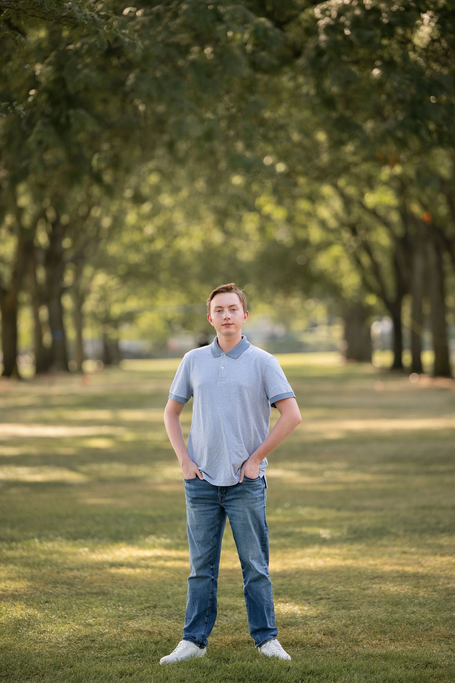 Young man in gray polo and jeans stands confidently in a tree-lined park path on a sunny day.