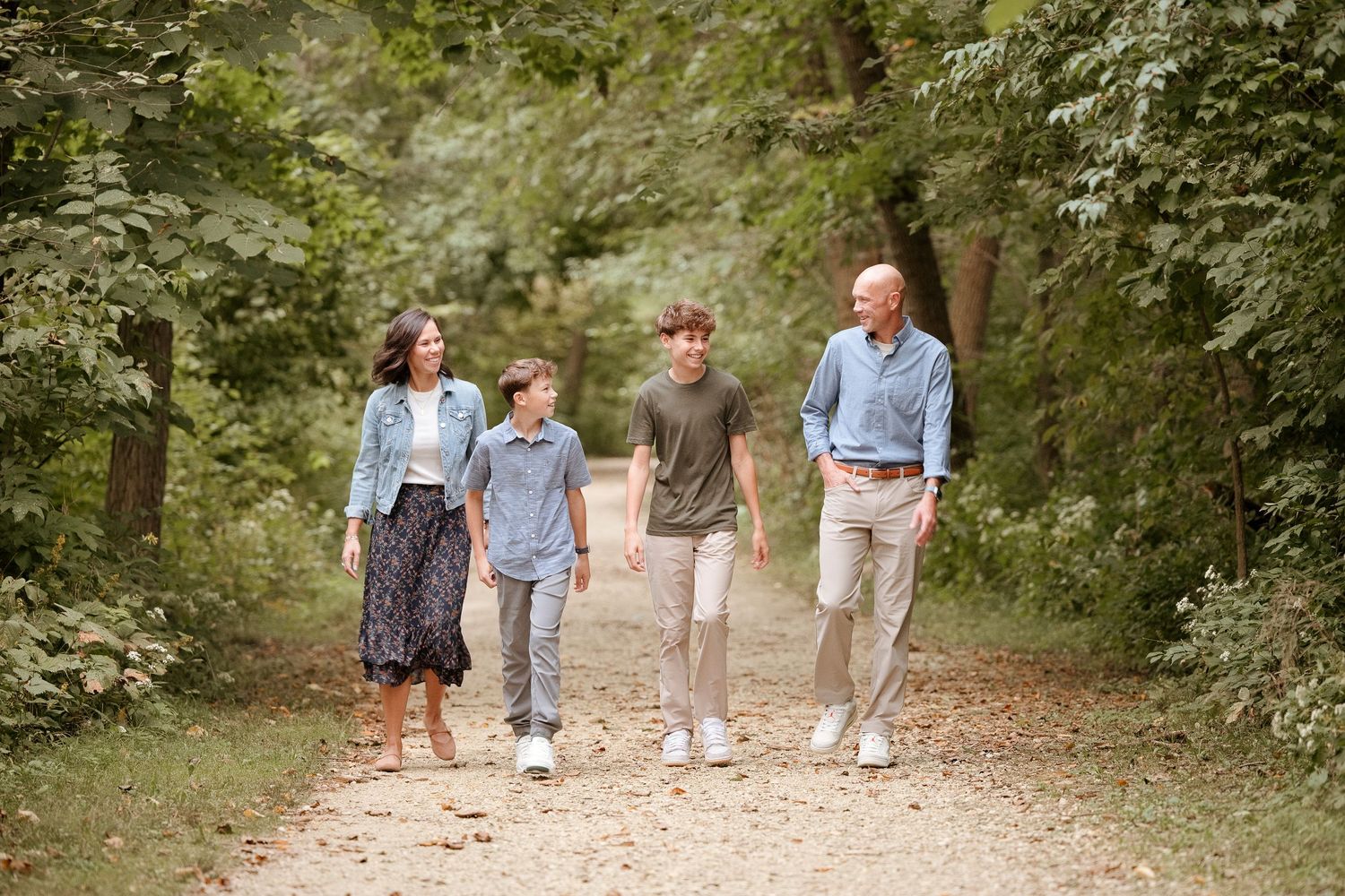 Family of four walking together on a scenic wooded path surrounded by lush green trees.