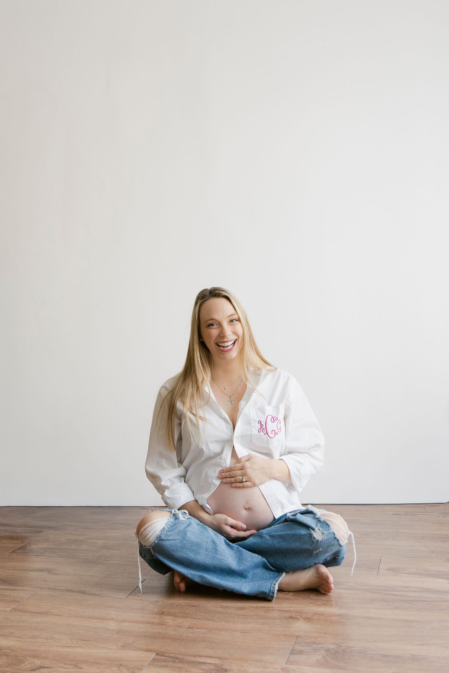 Expectant mother in white shirt and jeans sits cross-legged on wooden floor against white wall in maternity photoshoot.
