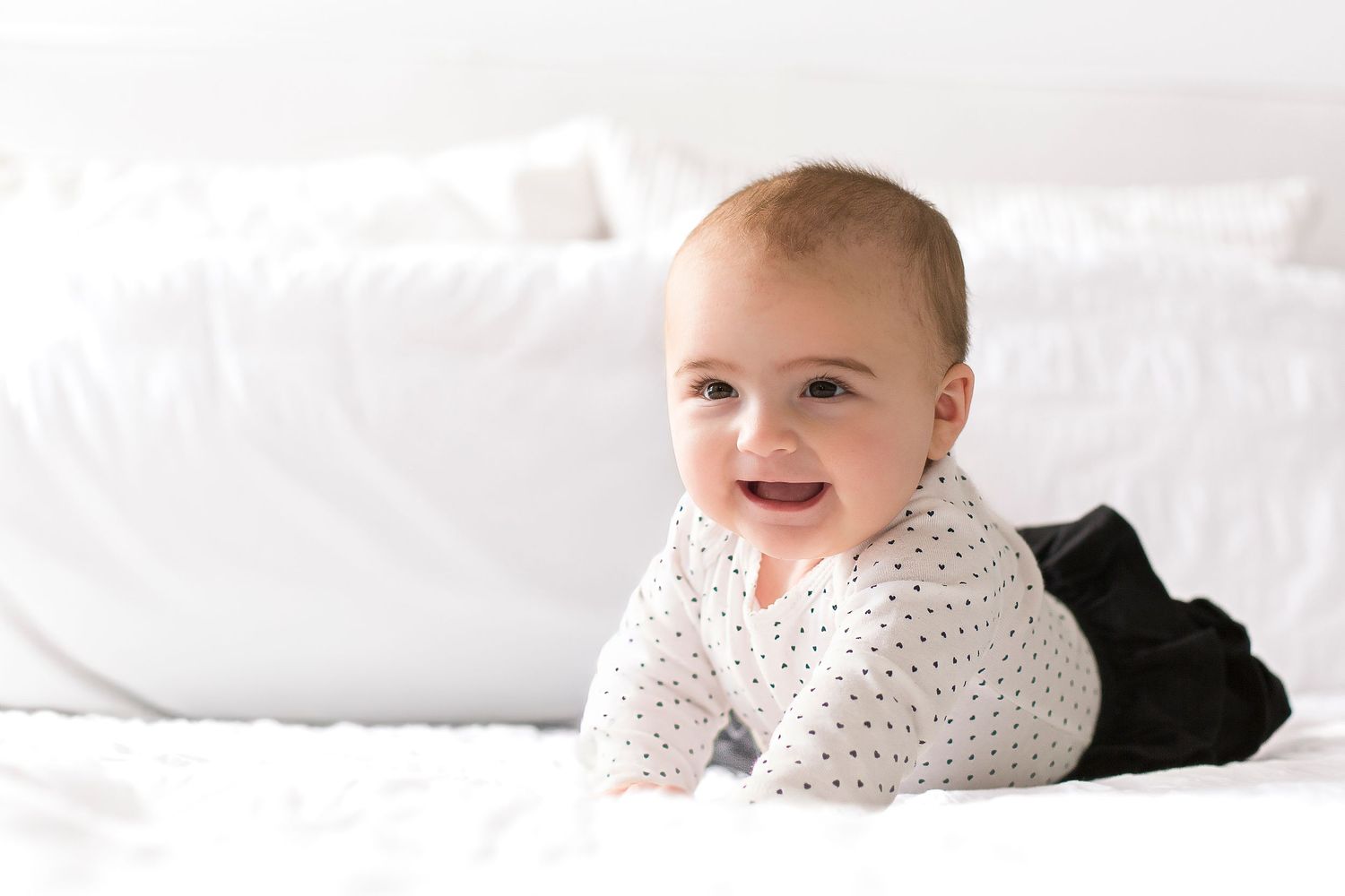 Baby in polka dot outfit lying on white bedding while looking to the side with a sweet expression.