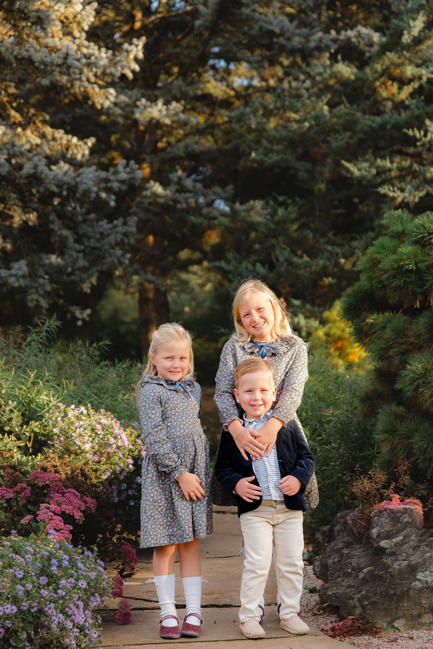 A family poses together on a garden path surrounded by evergreen trees and pink flowers in warm evening light.