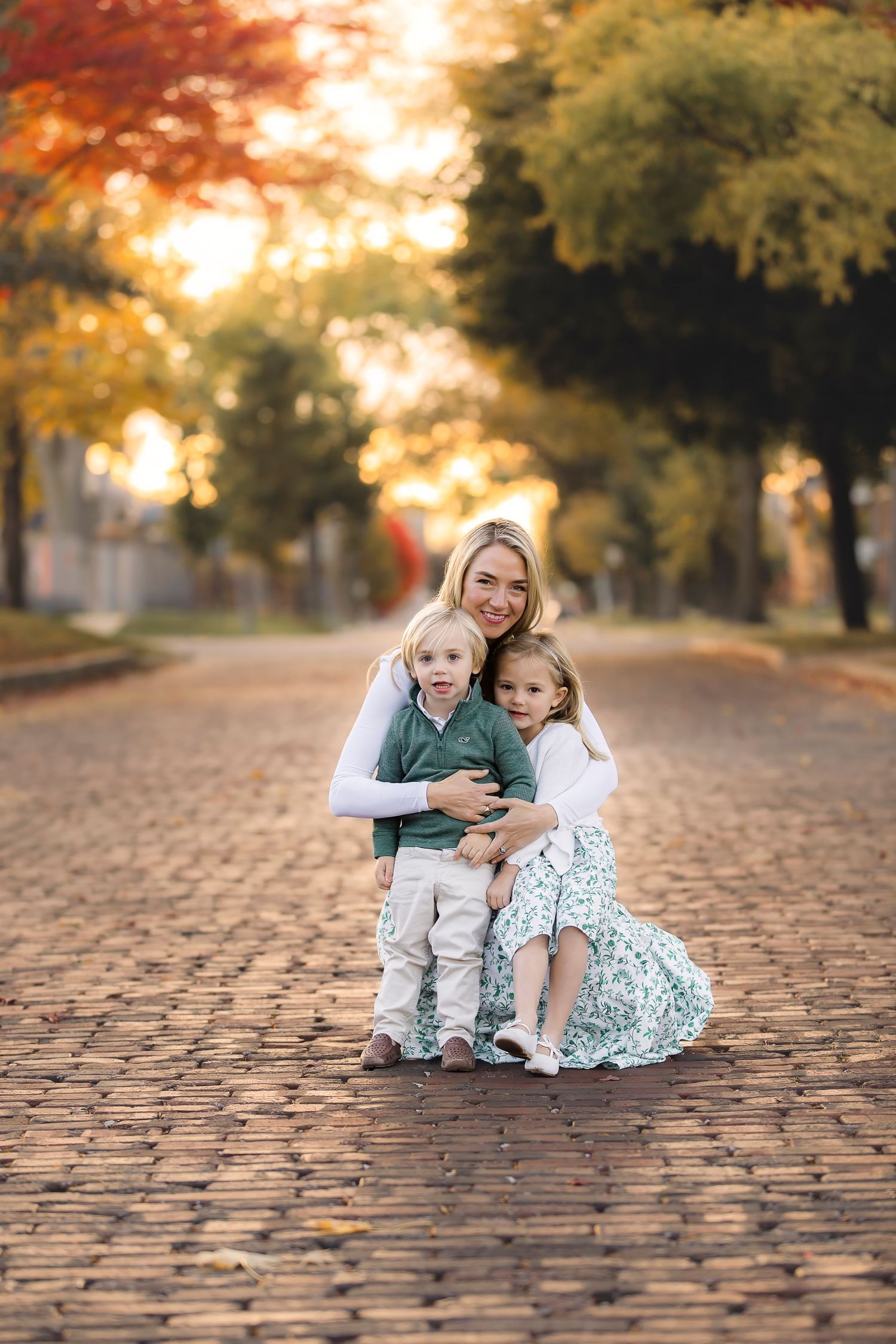 A family poses together on a cobblestone path during autumn with golden trees and sunlight in the background.