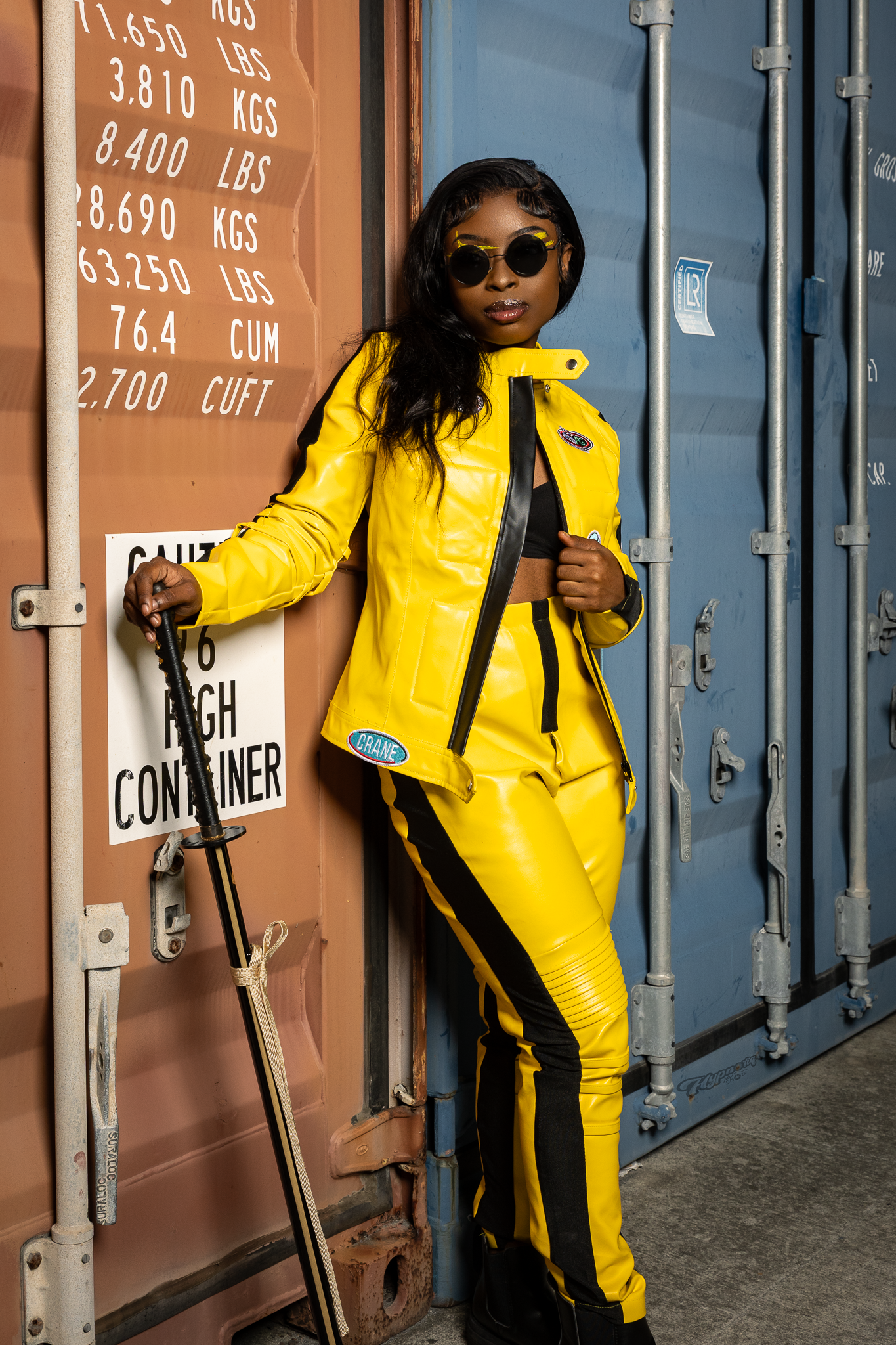 Woman in yellow leather racing suit and sunglasses posing against shipping containers.