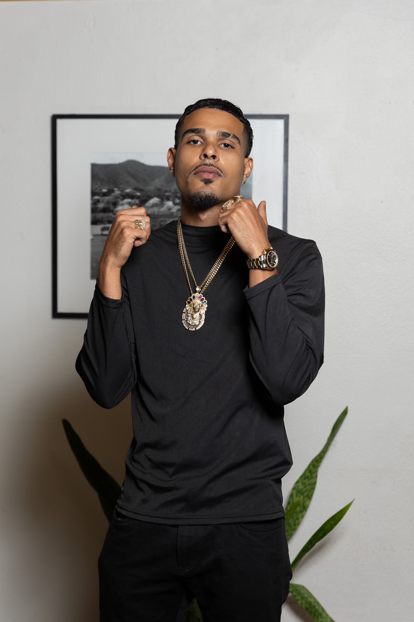 Young man in black outfit wearing gold chain necklace and watch, posing confidently indoors near a plant and framed photo.