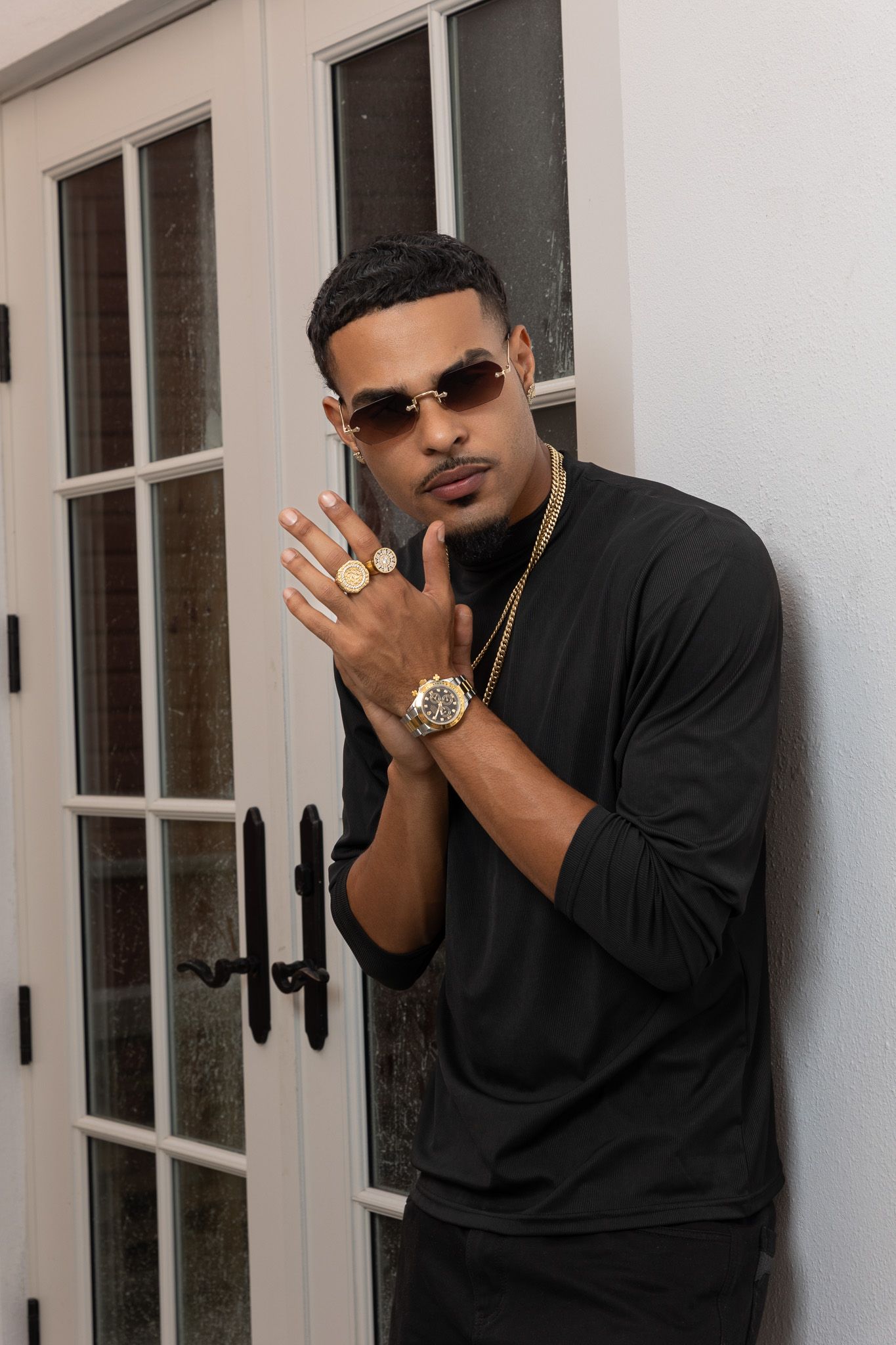 Young man in black outfit poses with gold jewelry, watch, rings, and sunglasses near white French doors.