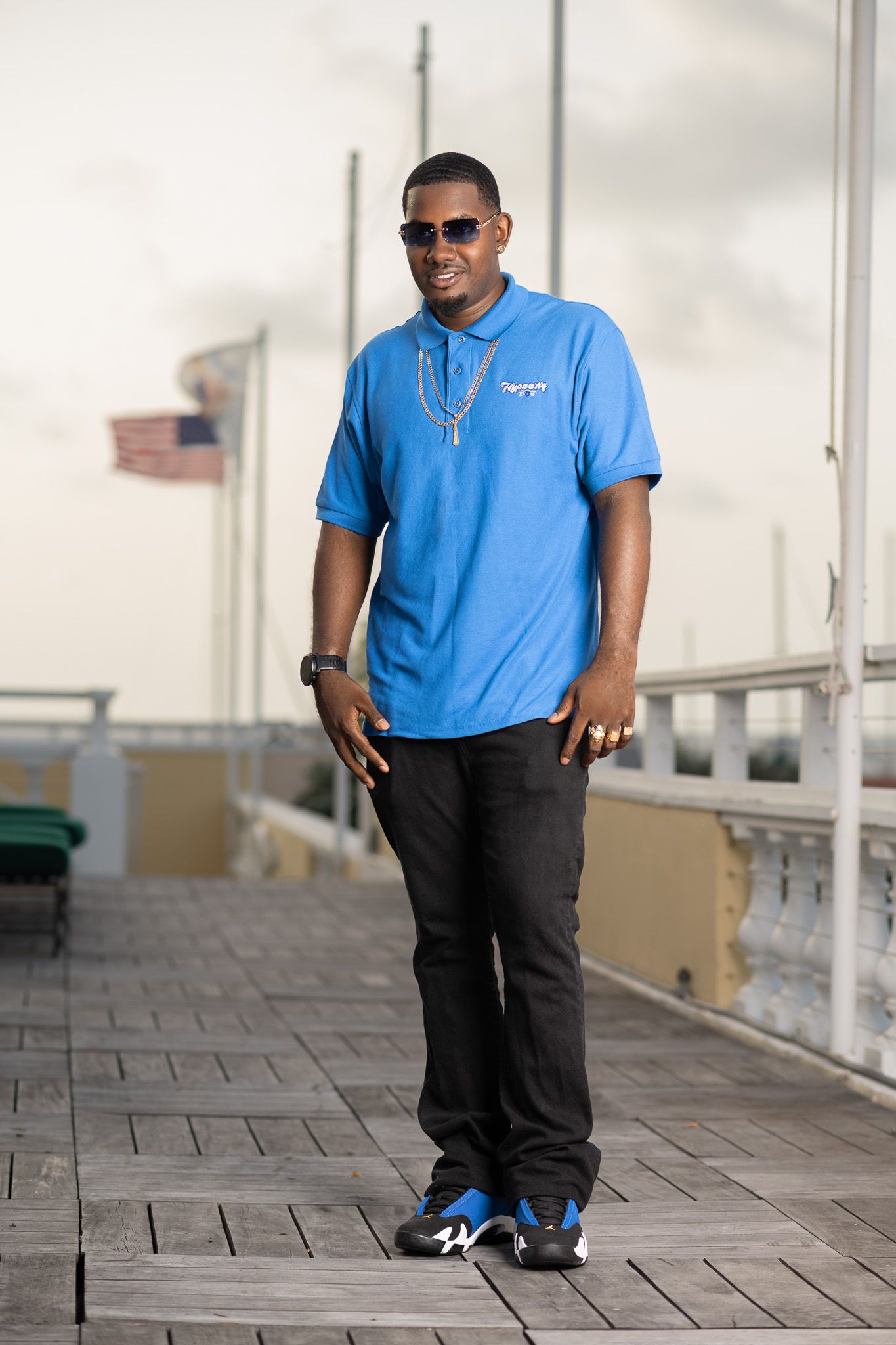 Smiling man in blue polo shirt, black jeans, and sneakers standing on a boardwalk with an American flag in background.