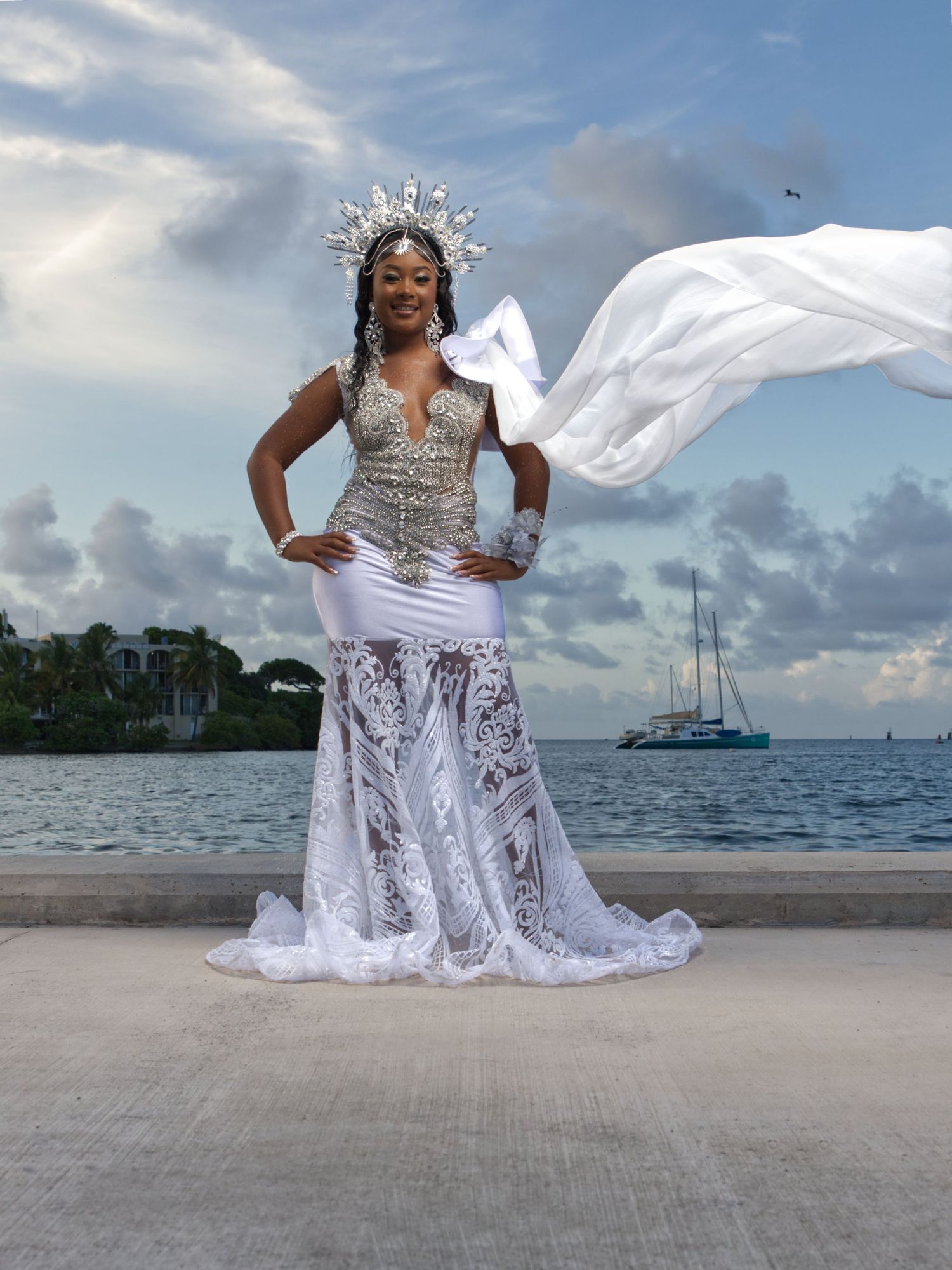 White lace dress and crown photographed on a beach with boats in the background.