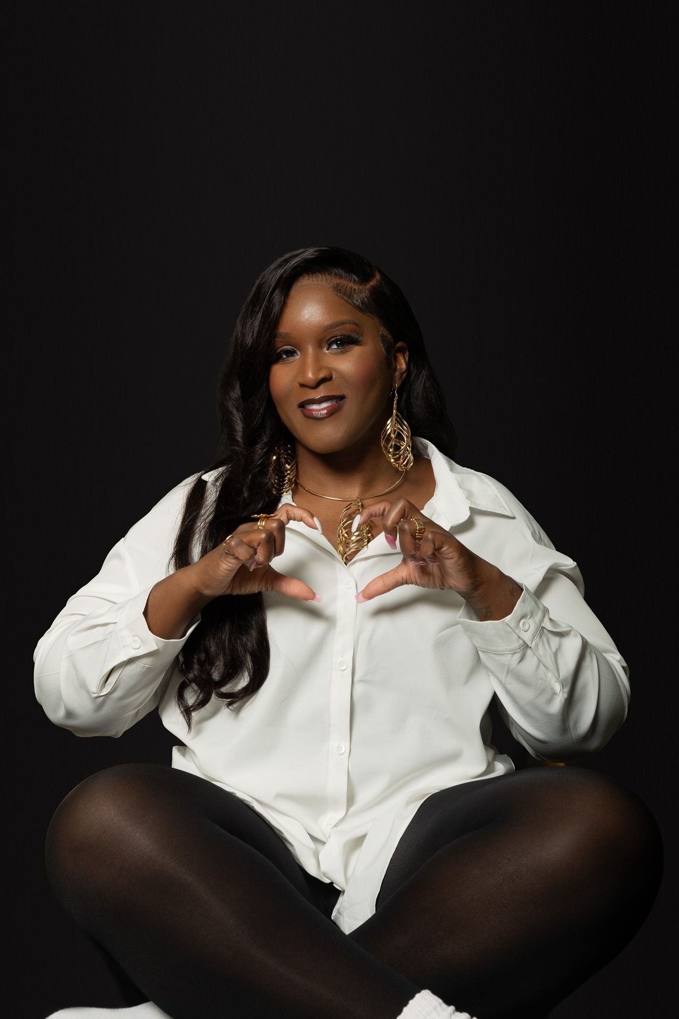 A smiling Black woman sits cross-legged wearing a white shirt and gold jewelry against a dark background.