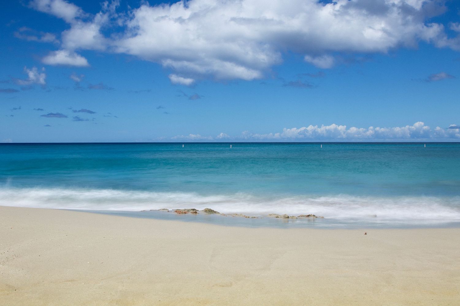 Pristine tropical beach landscape with turquoise waters meeting white sand under blue skies and fluffy clouds.