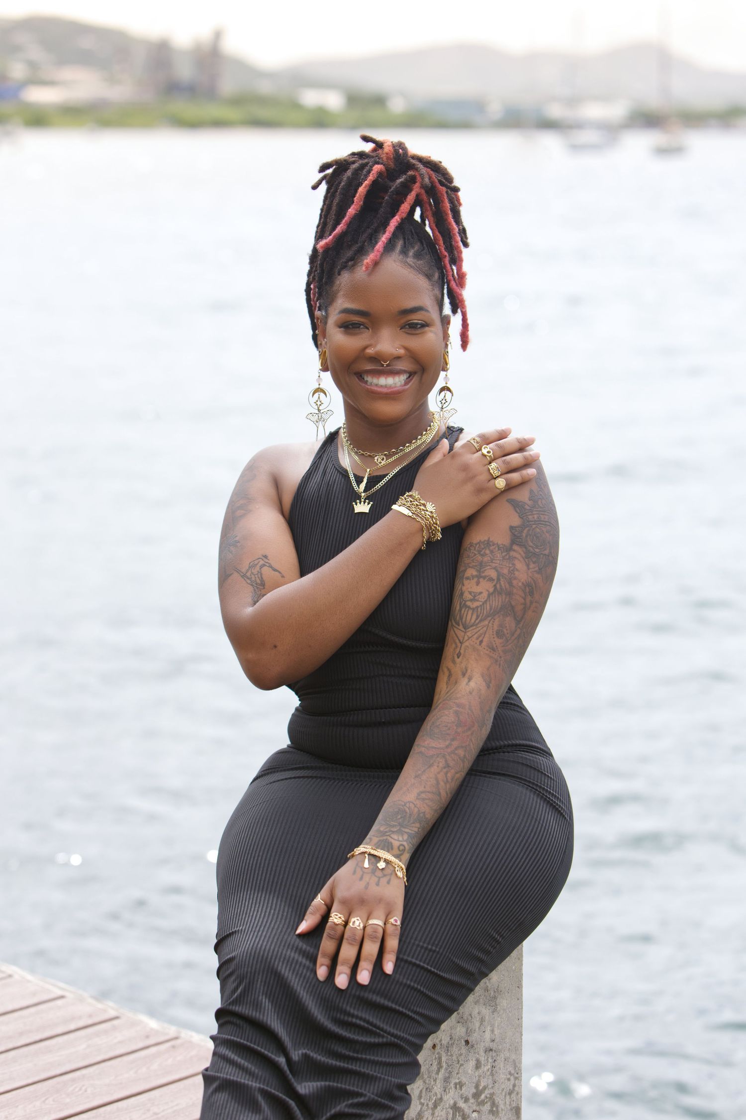 Casual outdoor photoshoot on wooden dock by waterfront showing tattoos and black outfit.