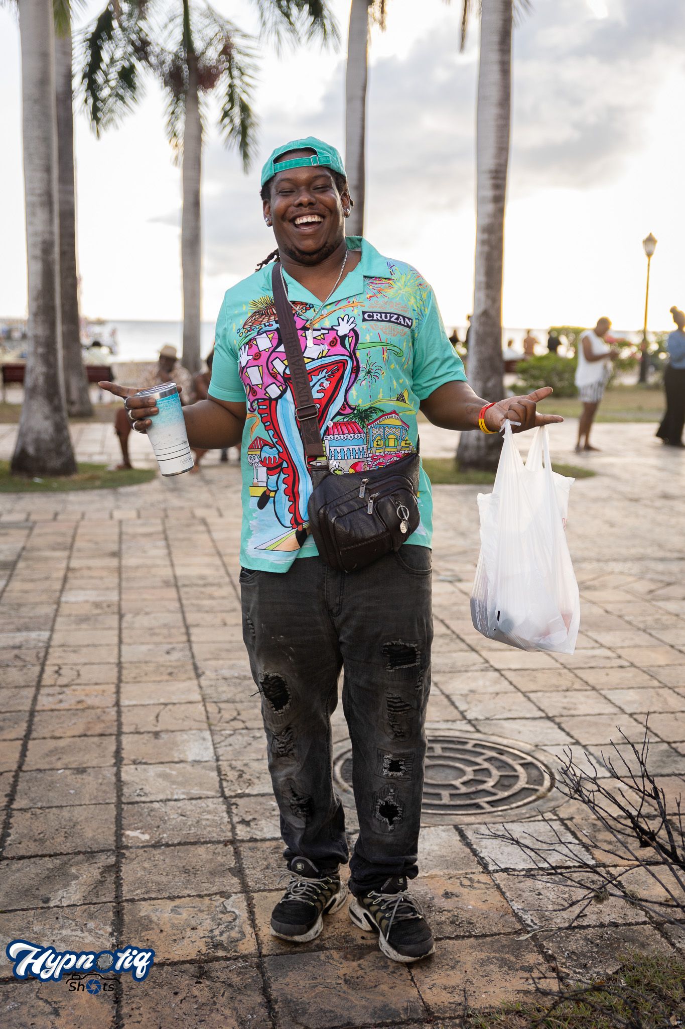 Person in teal shirt and cargo pants posing on a palm-lined walkway.