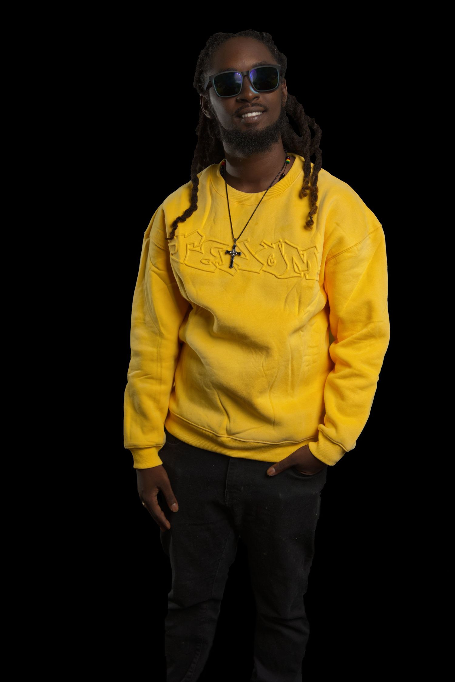 Fashion portrait of someone in yellow sweatshirt and dark pants wearing sunglasses against black backdrop.