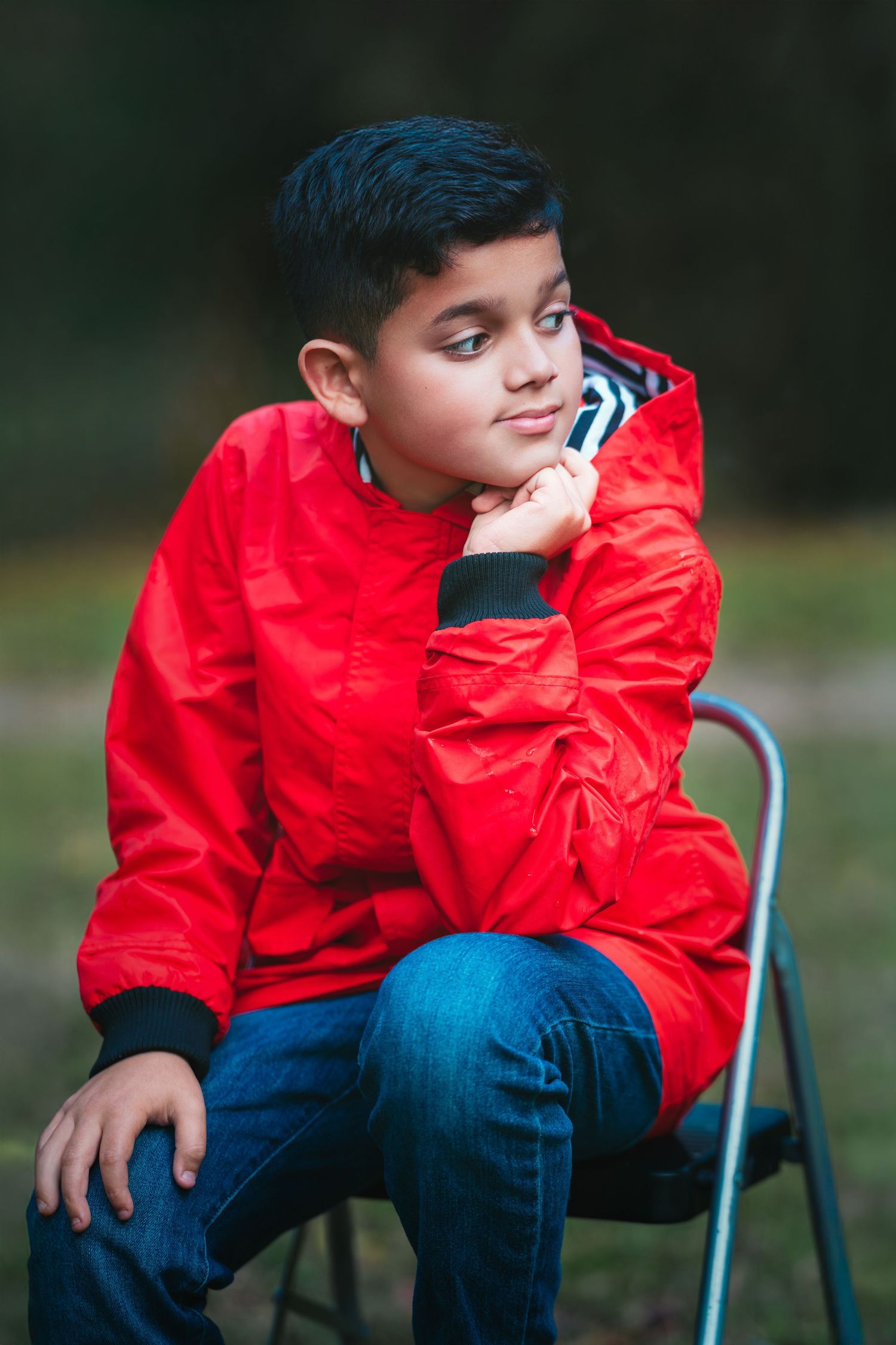 Child in red windbreaker and jeans sits thoughtfully on outdoor chair in natural setting.