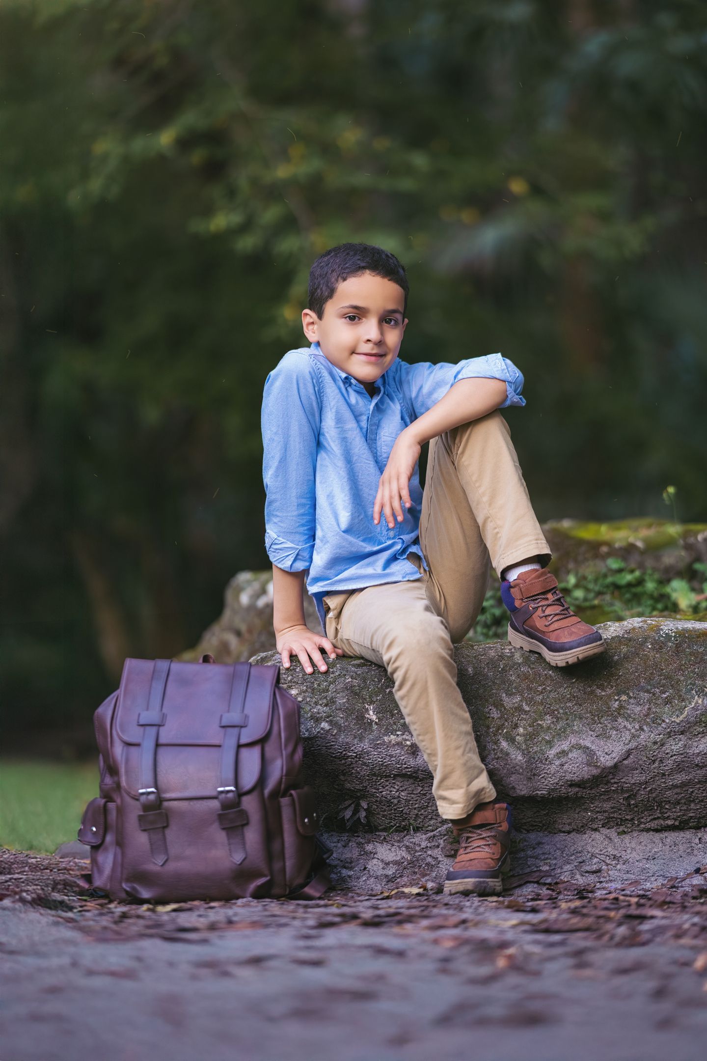 Boy in blue shirt and khaki pants sitting on a rock with a brown leather backpack in a park.
