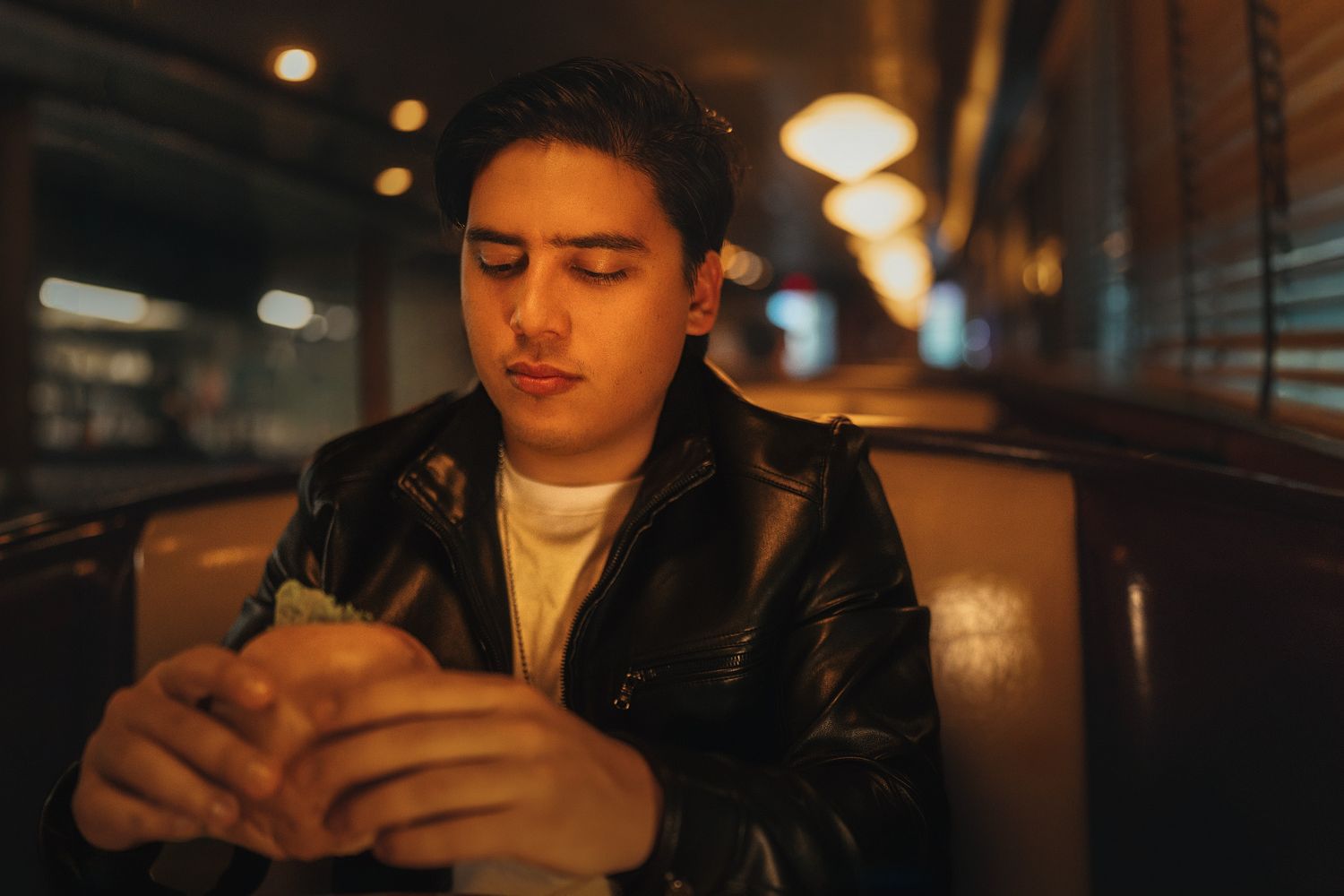 Young man in black leather jacket looking down at food in a cozy vintage diner booth.
