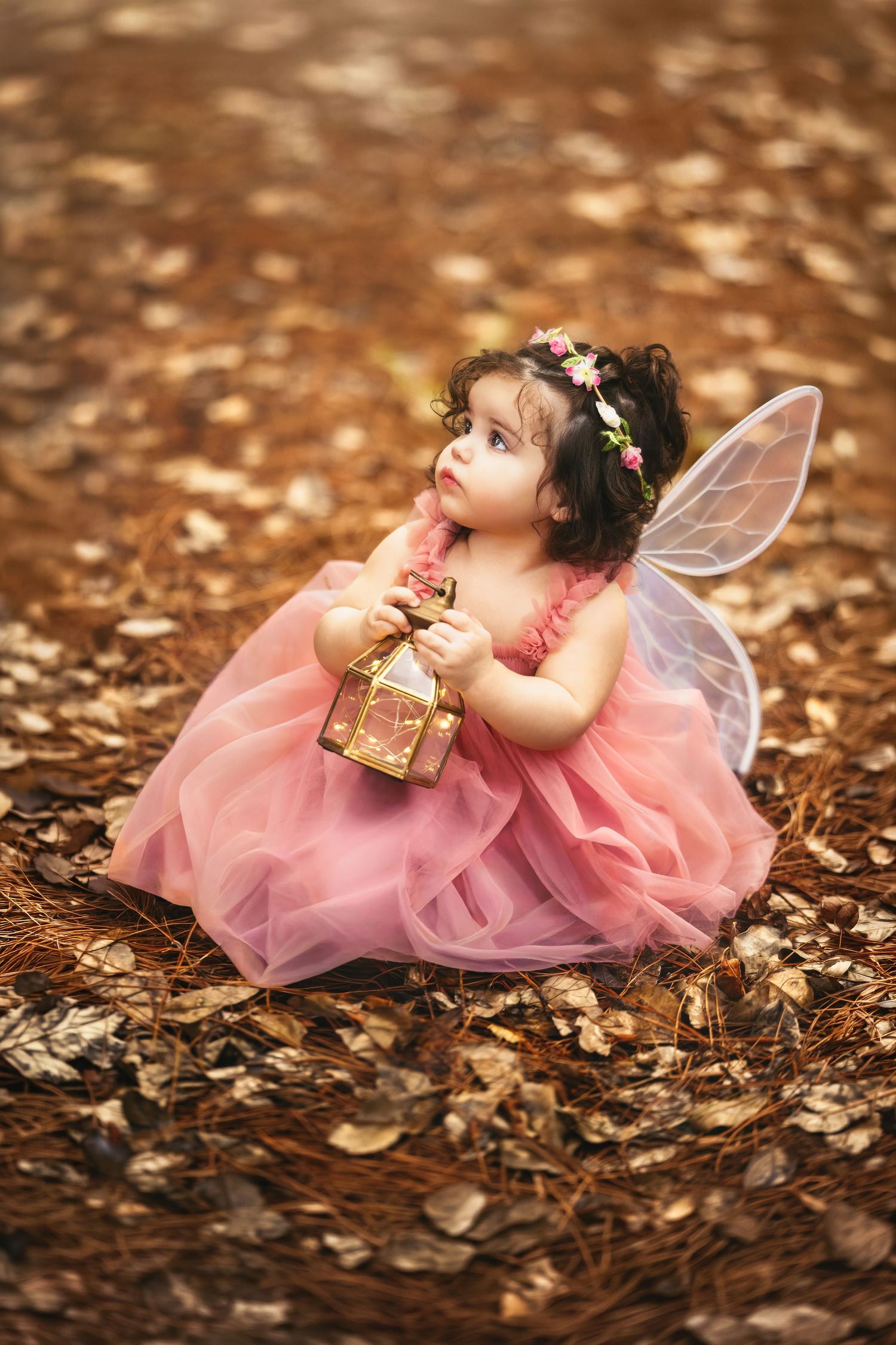 Toddler girl in pink tutu dress with fairy wings and floral crown holds lantern outdoors on autumn leaves.